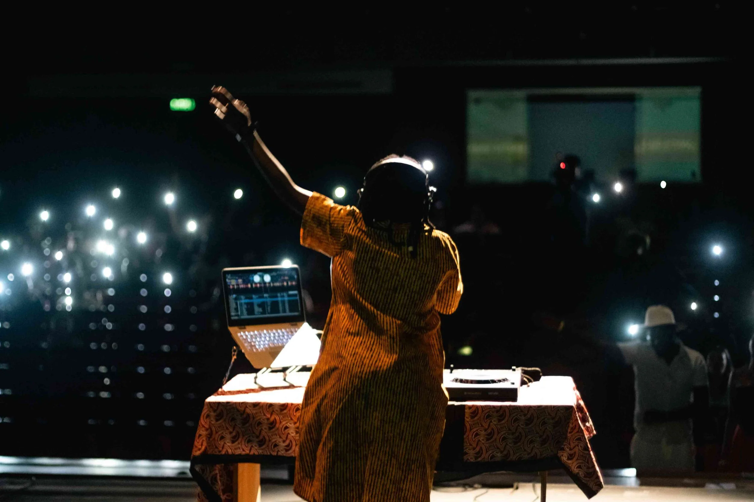 A DJ wearing a yellow striped shirt and helmet performing at a dark event with her arms raised, surrounded by an audience with many illuminated by phone lights.