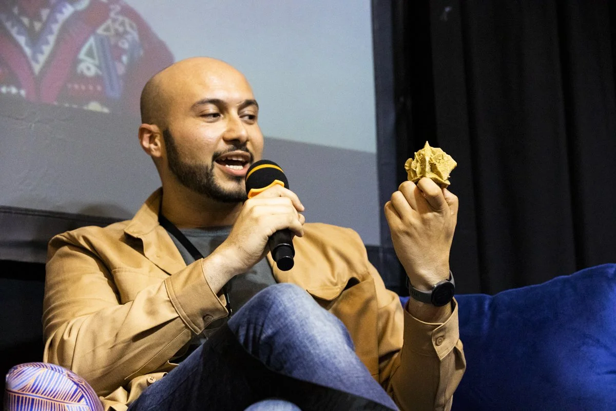 A man with a beard and mustache, holding a microphone in one hand and a crumpled cloth or paper in the other, sitting and speaking in front of a projection screen.