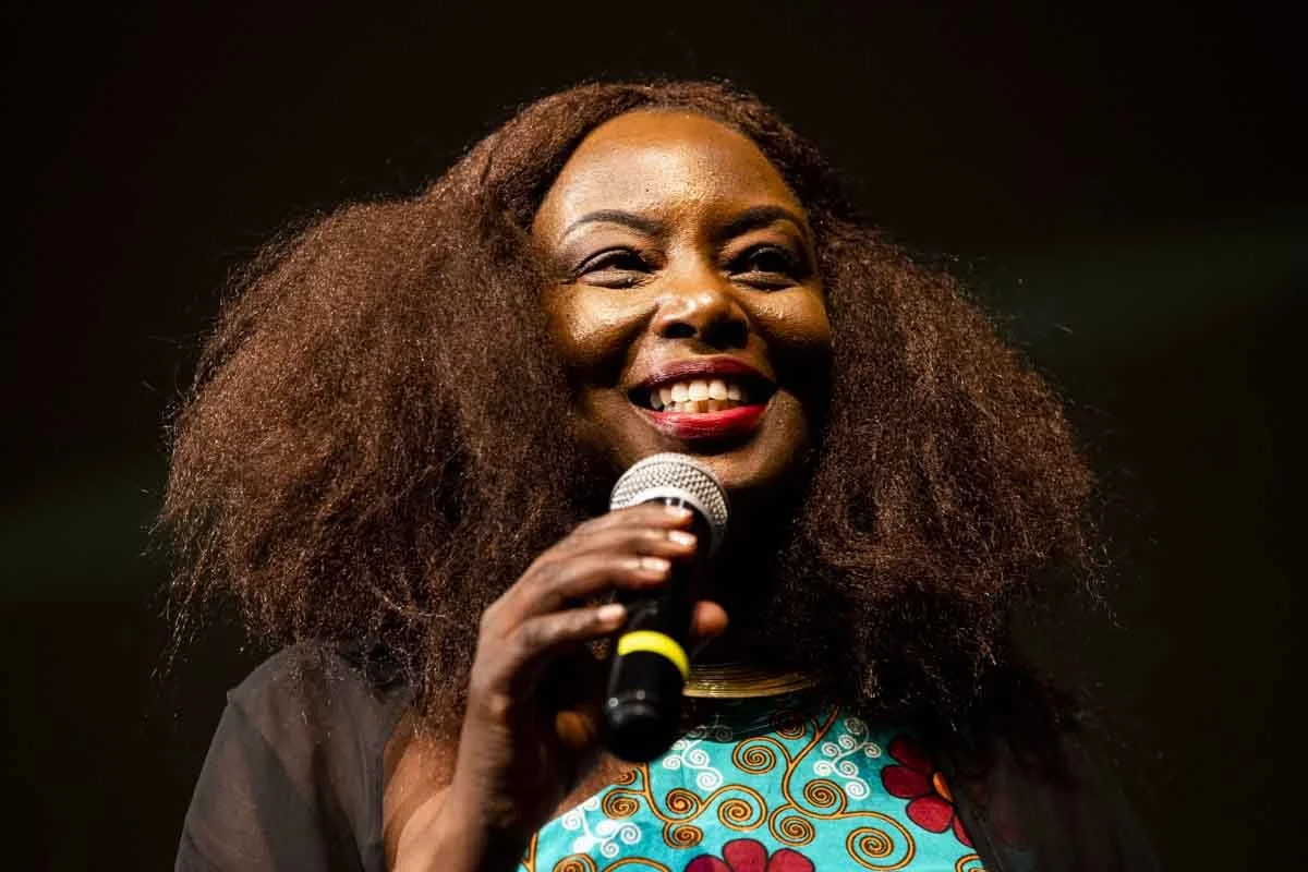 A woman with curly hair smiling and holding a microphone during a speech or performance.