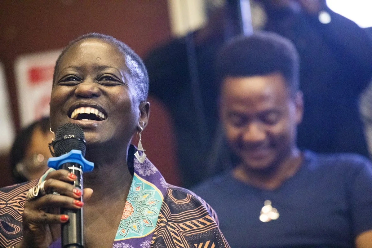 A woman with short gray hair smiling and holding a microphone, wearing a colorful patterned outfit and earrings, with a woman in the background looking down.