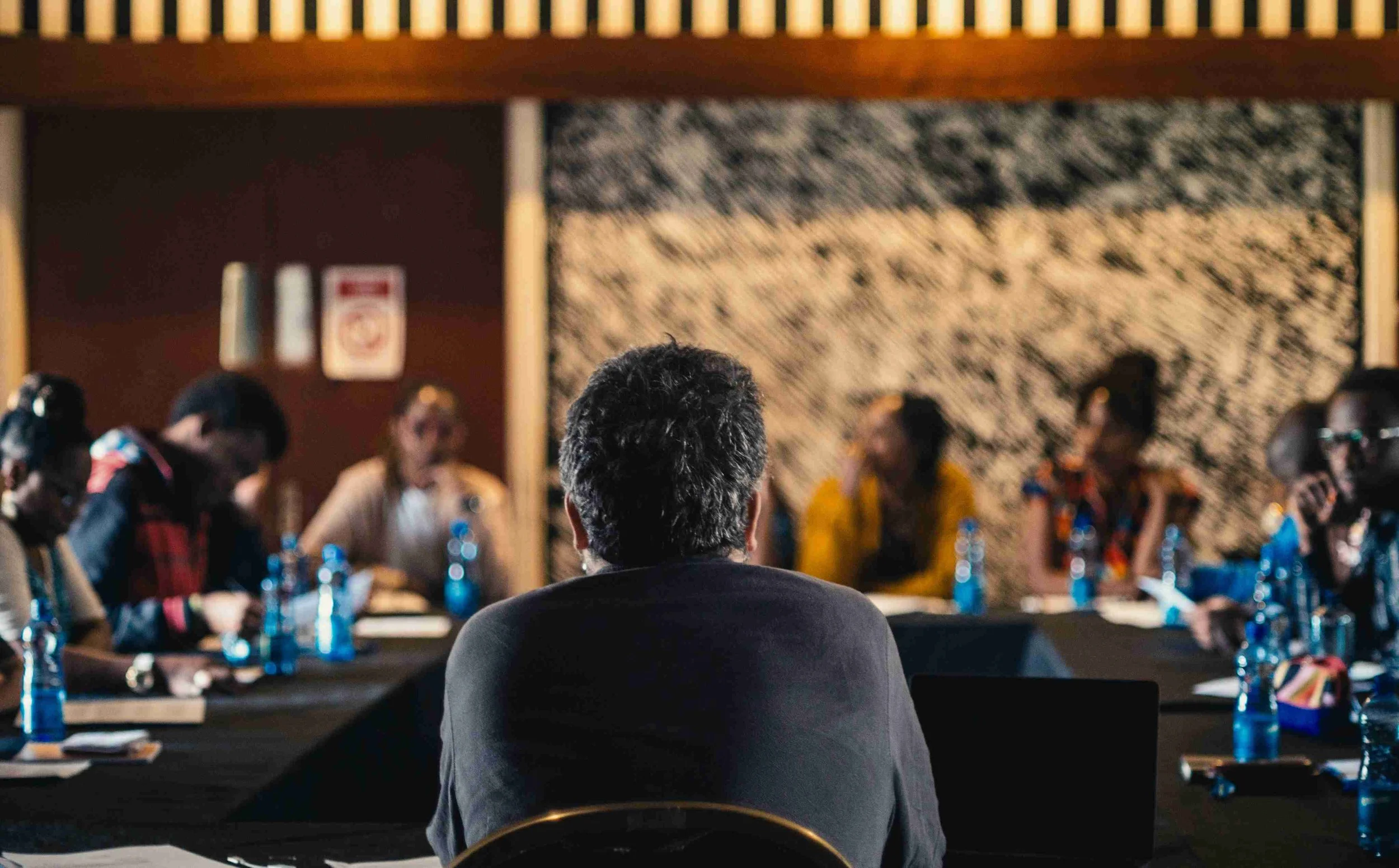 A person with gray hair sits at a conference table facing a panel of people in a dimly lit room.