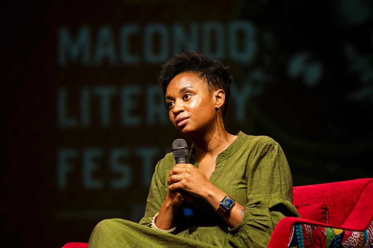 A woman with short black curly hair, wearing a green dress and a smartwatch, sitting on a red chair with a colorful pattern, holding a microphone, speaking at an event with a background that says 'Macondo Literary Festival.'