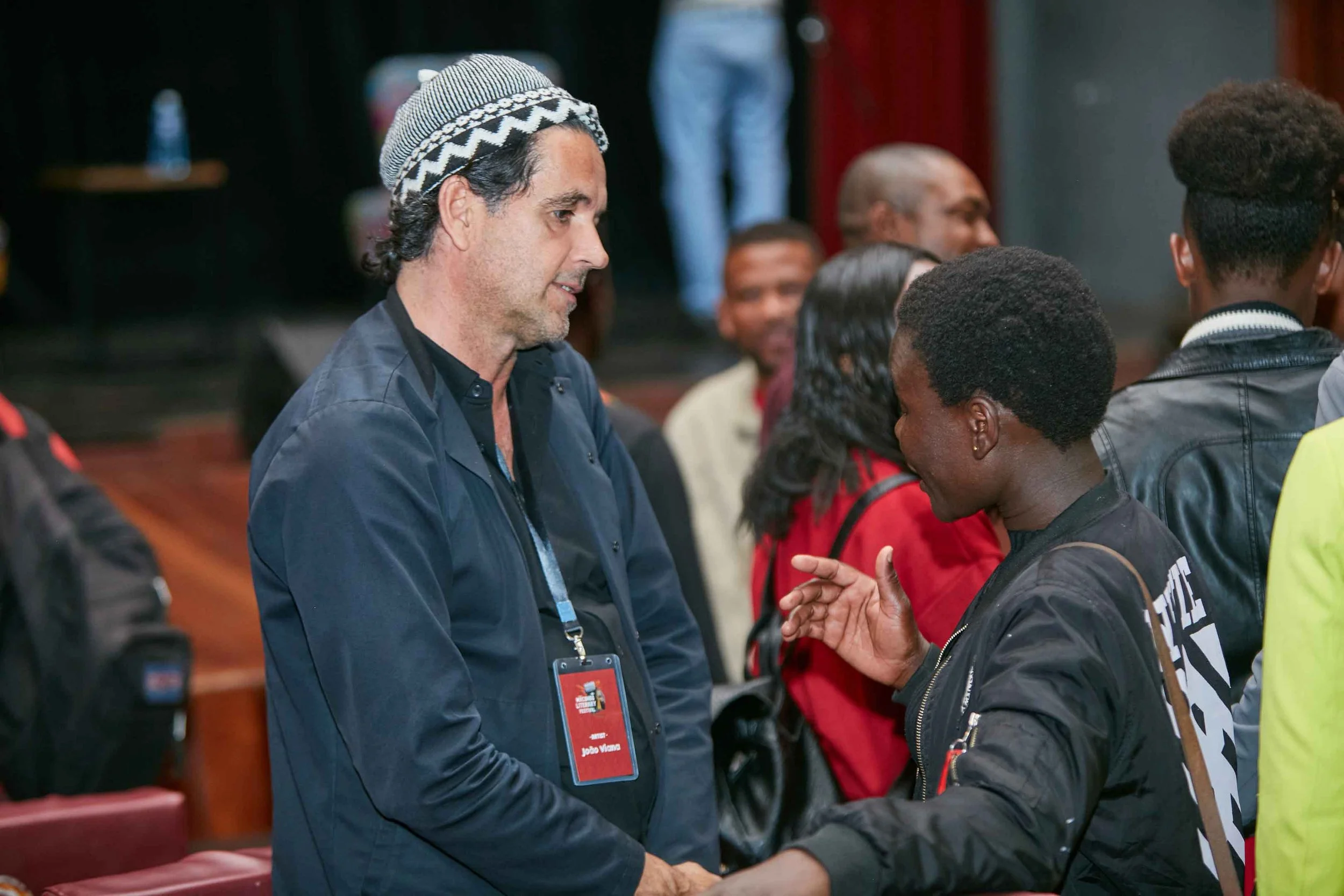 Two men shaking hands at an indoor event, with several other people engaged in conversation in the background.