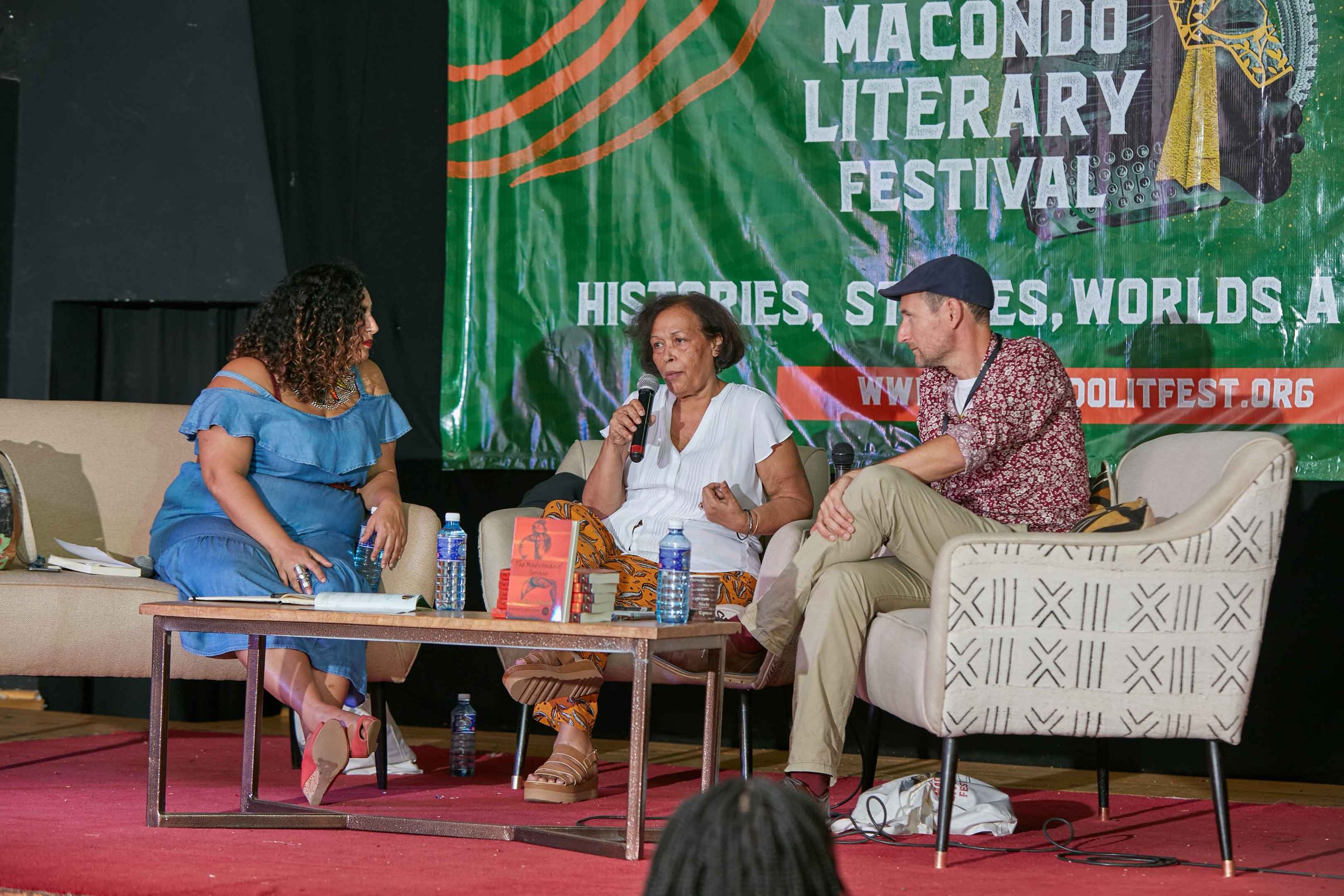 Three people sitting on stage at Macondo Literary Festival, engaging in a discussion. The woman in the middle is speaking into a microphone, flanked by a woman on the left in a blue dress and a man on the right in a patterned shirt and cap. A large b