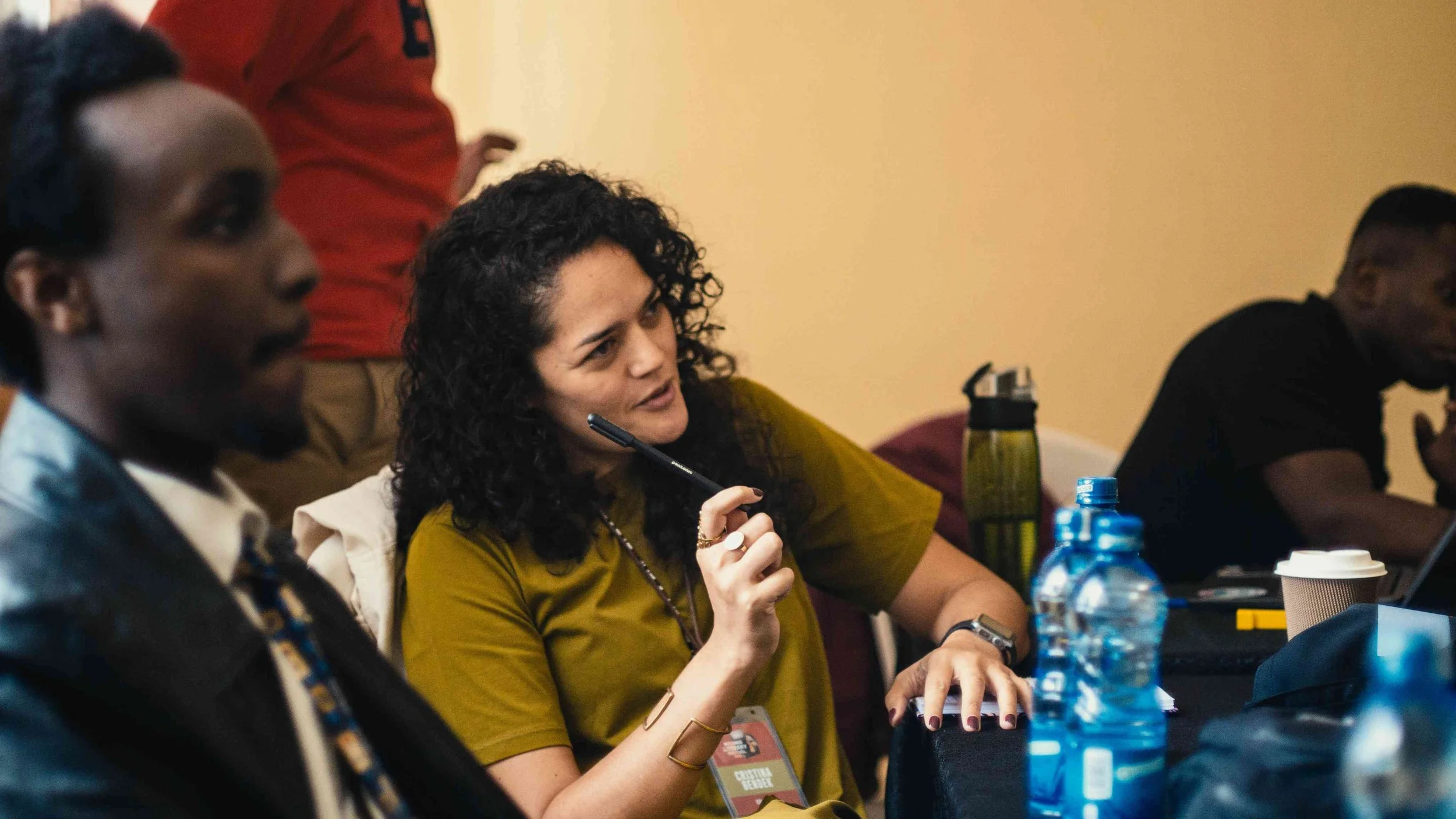 A woman with curly dark hair, wearing an olive green shirt, is sitting at a table with a pen in her hand, engaging in a discussion or meeting. Beside her, there are water bottles and a coffee cup on the table. Other people are also present, some part