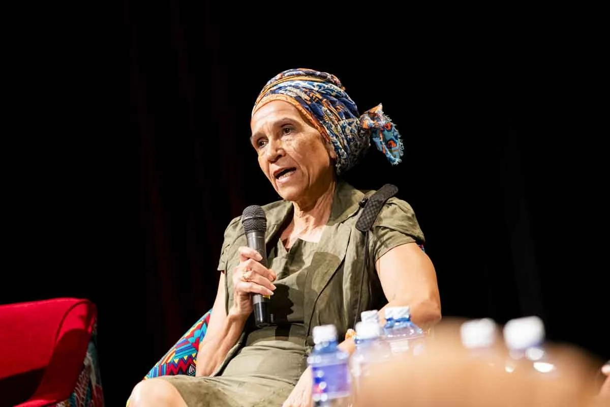 An elderly woman speaking into a microphone during a discussion or interview on a stage, with water bottles in front of her and a dark background.