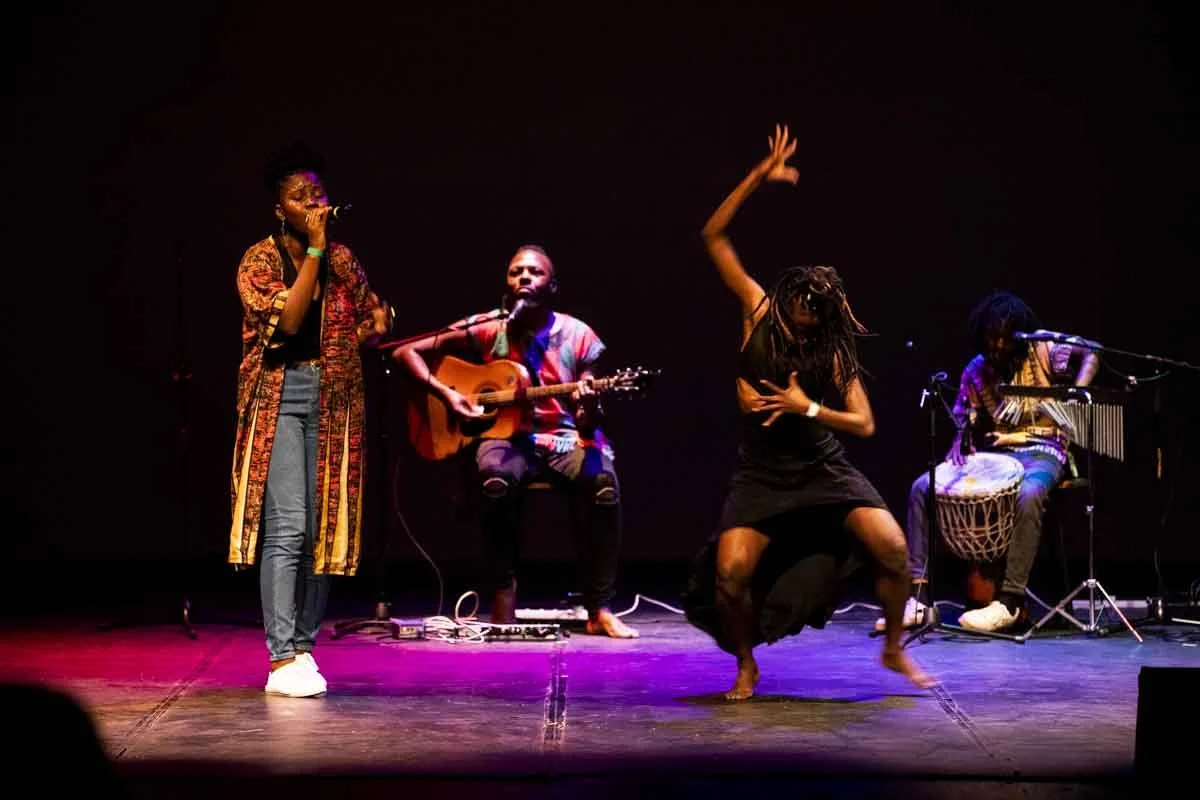 Four performers on stage, one singing into a microphone, one playing guitar, one dancing with dreadlocks, and one playing drums, in a dark setting with stage lighting.