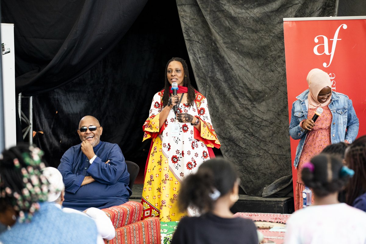 Three people on stage during an event, with women holding microphones and a seated man wearing sunglasses, audience members in front.