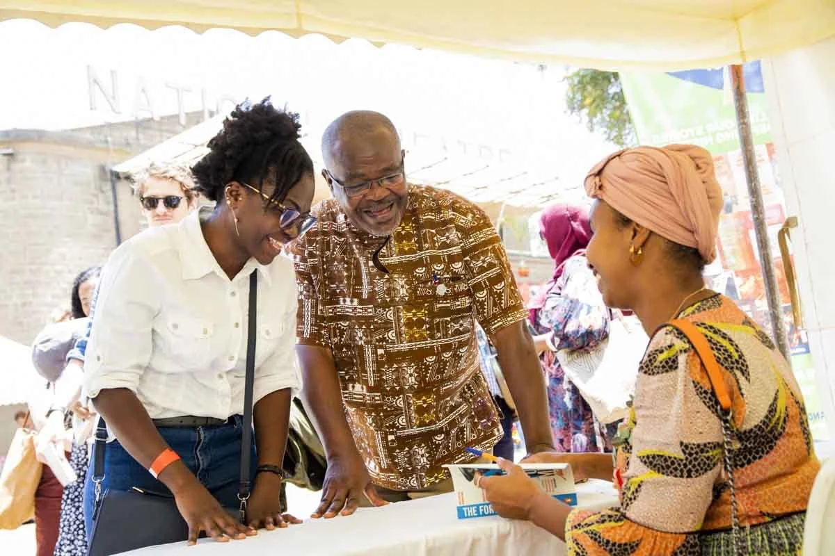 Three people smiling and engaging in conversation at an outdoor event under a tent. A woman wearing a pink headwrap is holding a brochure, while a young woman with glasses and a white shirt and an older man with glasses and a patterned shirt are lean