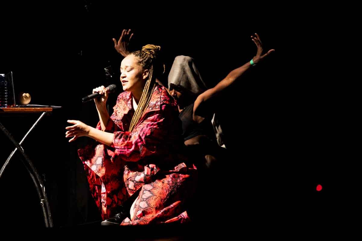 Two performers on stage, one singing into a microphone with eyes closed, wearing a red patterned robe, and the other with a hooded cloak and arms raised, set against a dark background.