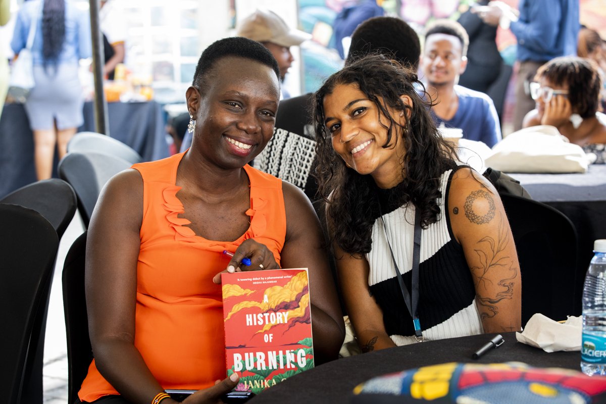 Two women smiling at a table during a book event, one holding a book titled "A History of Burning".