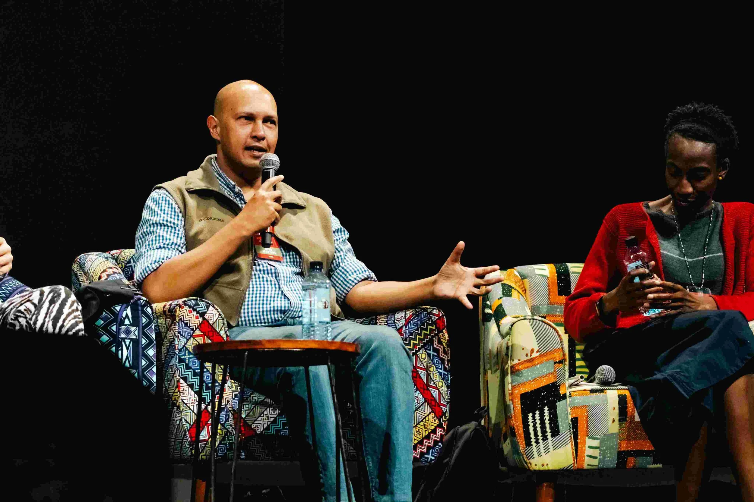 A man with a bald head and wearing a checkered shirt with a beige vest, speaking into a microphone on stage. A woman with short hair, wearing a red cardigan and black skirt, sits beside him, looking at her phone and holding a water bottle.