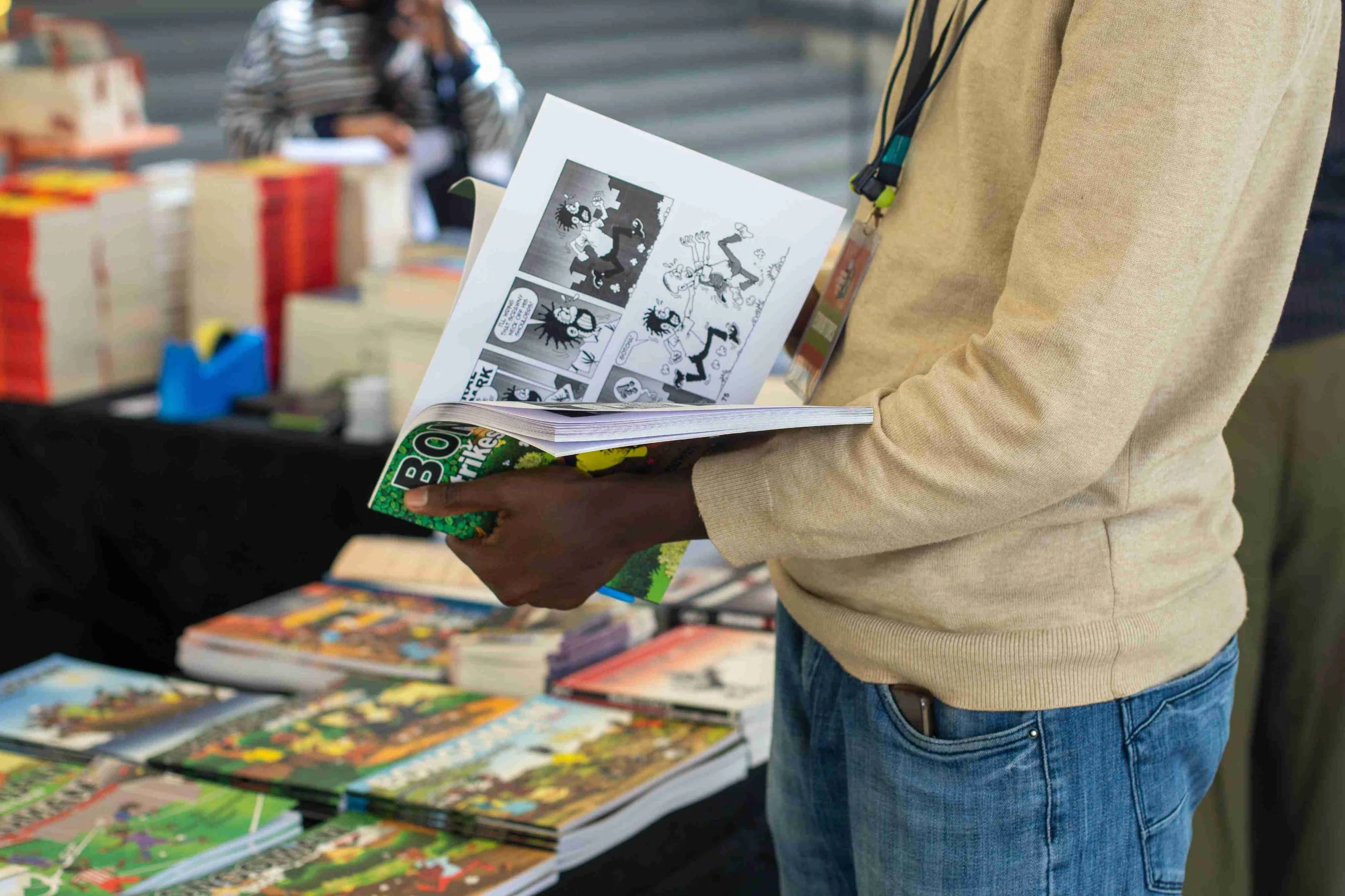 Person holding comic book and reading a cartoon comic strip at a book fair with various books on a table in the background.