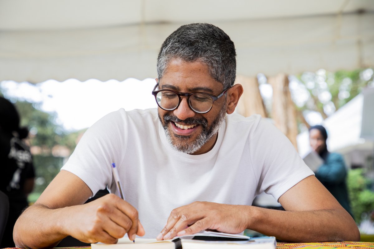 A middle-aged man with gray hair and glasses smiling while writing in a notebook at an outdoor event.