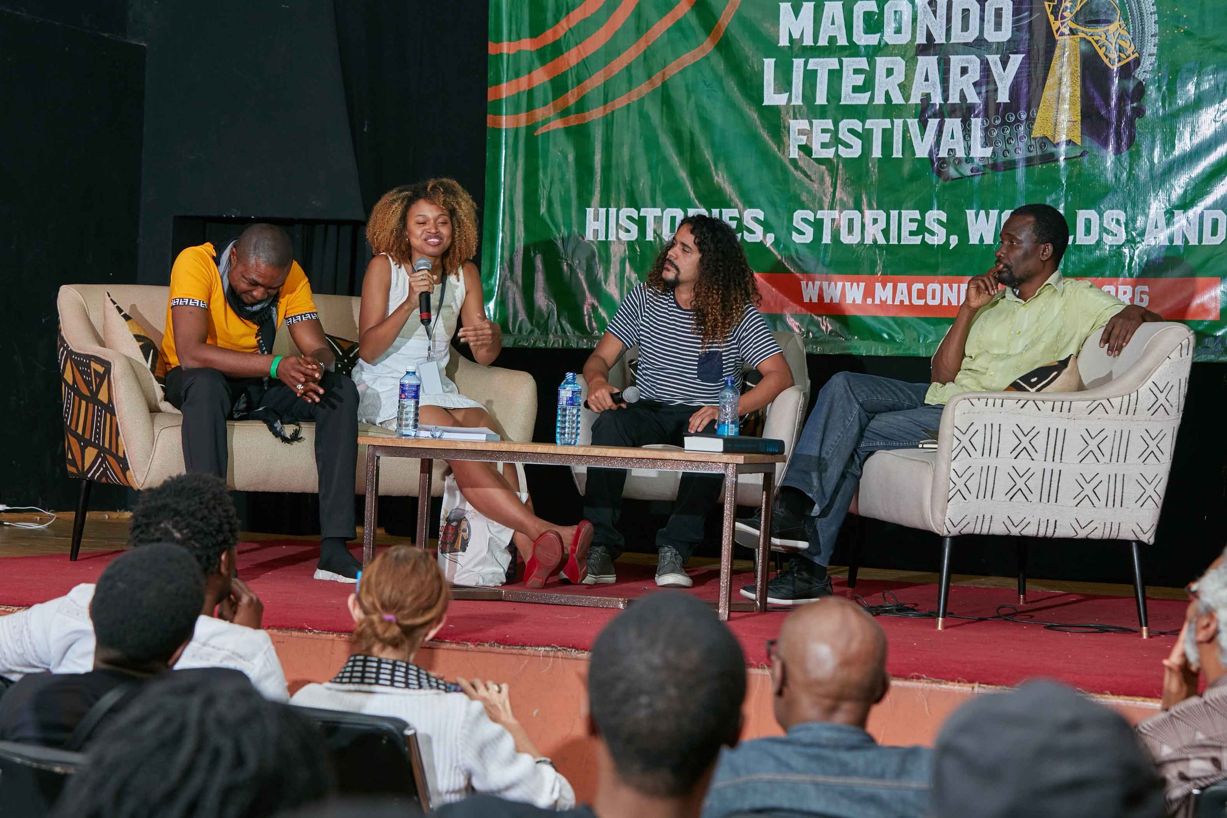 A panel discussion at the Macondo Literary Festival, with four people sitting on stage and engaging in conversation. An audience is visible in front of the stage. The background features a large green banner with the event's name and theme.