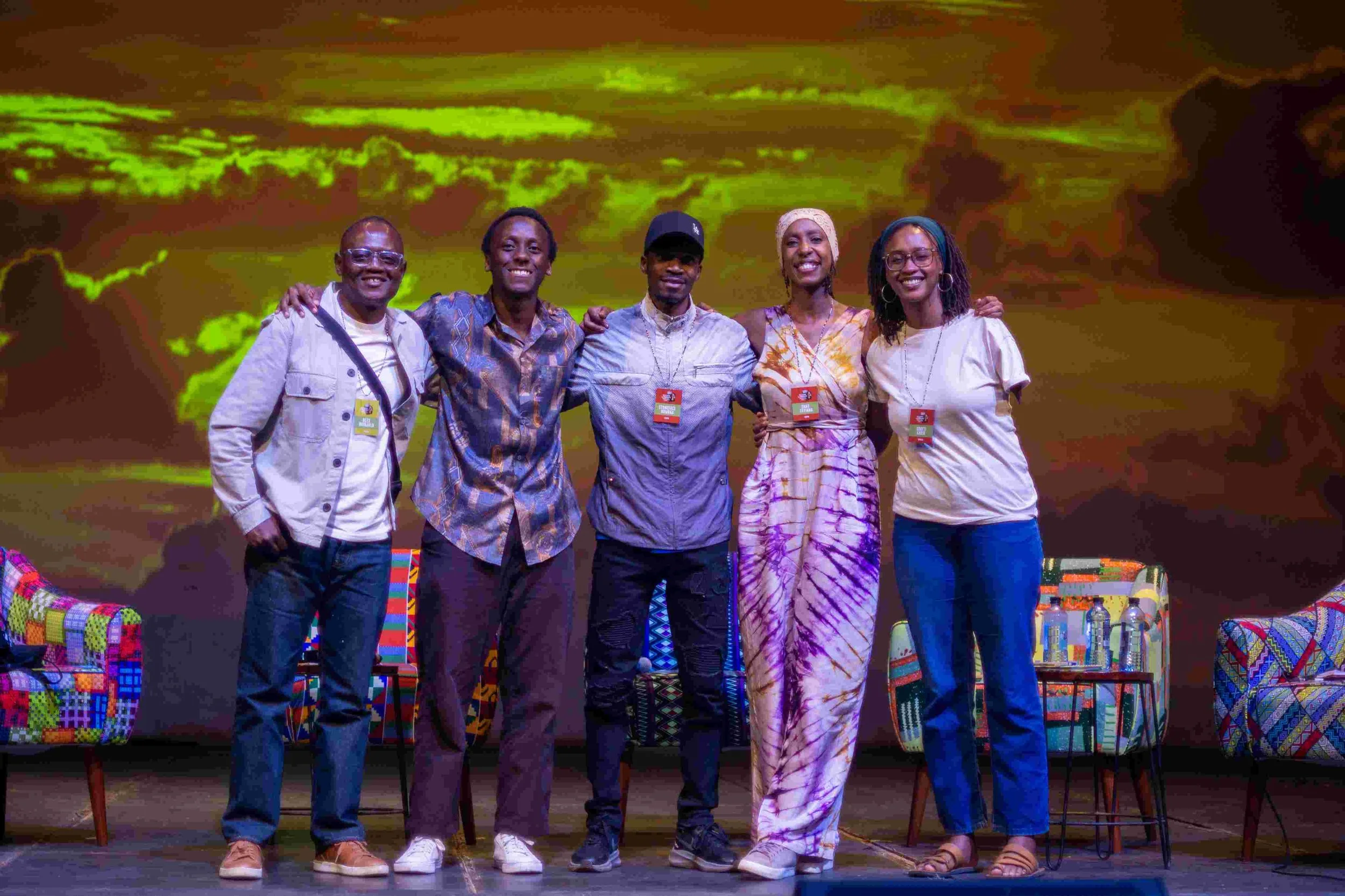 A group of five diverse people standing together on a stage, smiling, with colorful chairs and a vibrant, illuminated background featuring swirling clouds.