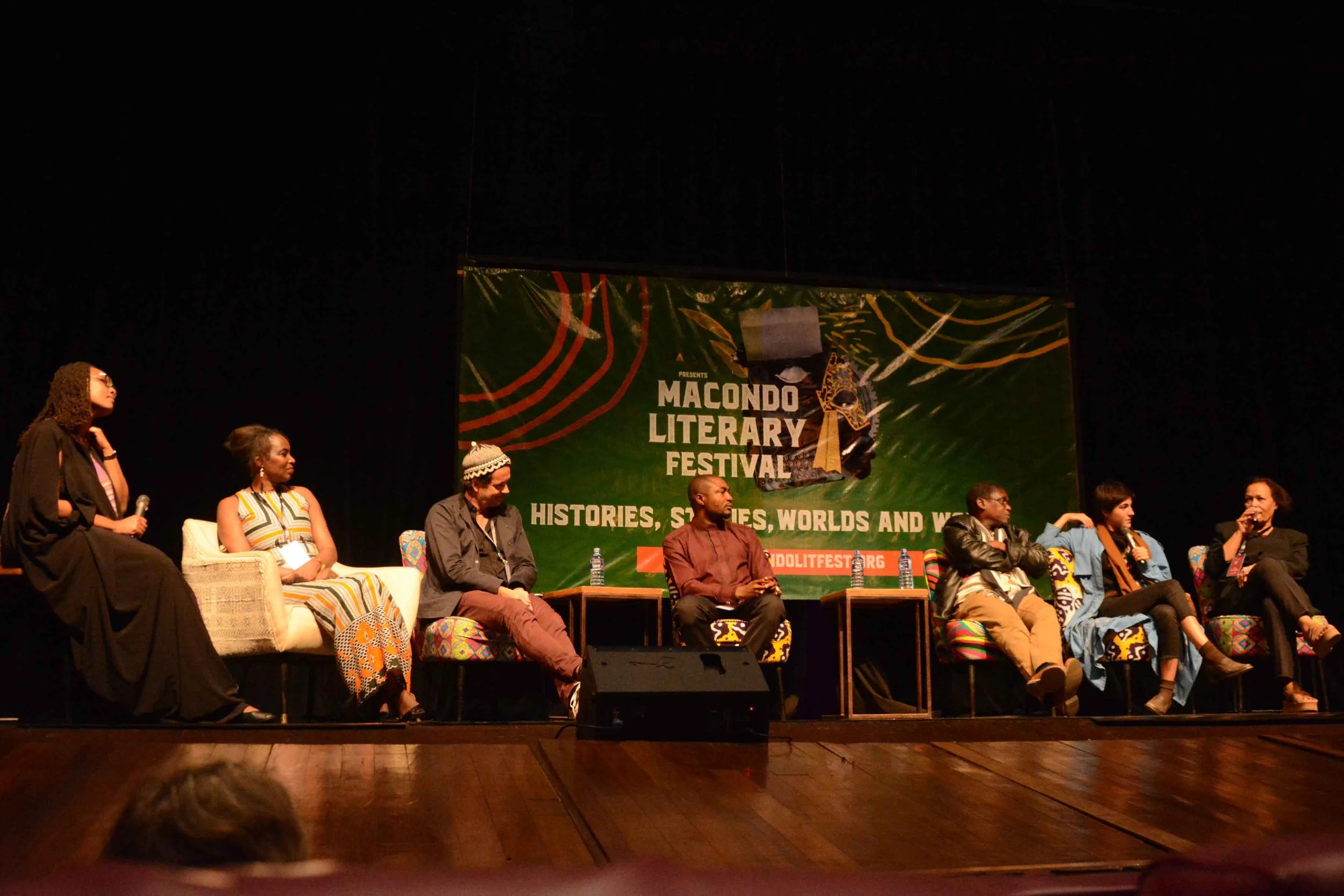 Panel discussion at the Macondo Literary Festival with seven individuals seated on stage, singing, and chatting, behind a large green banner.