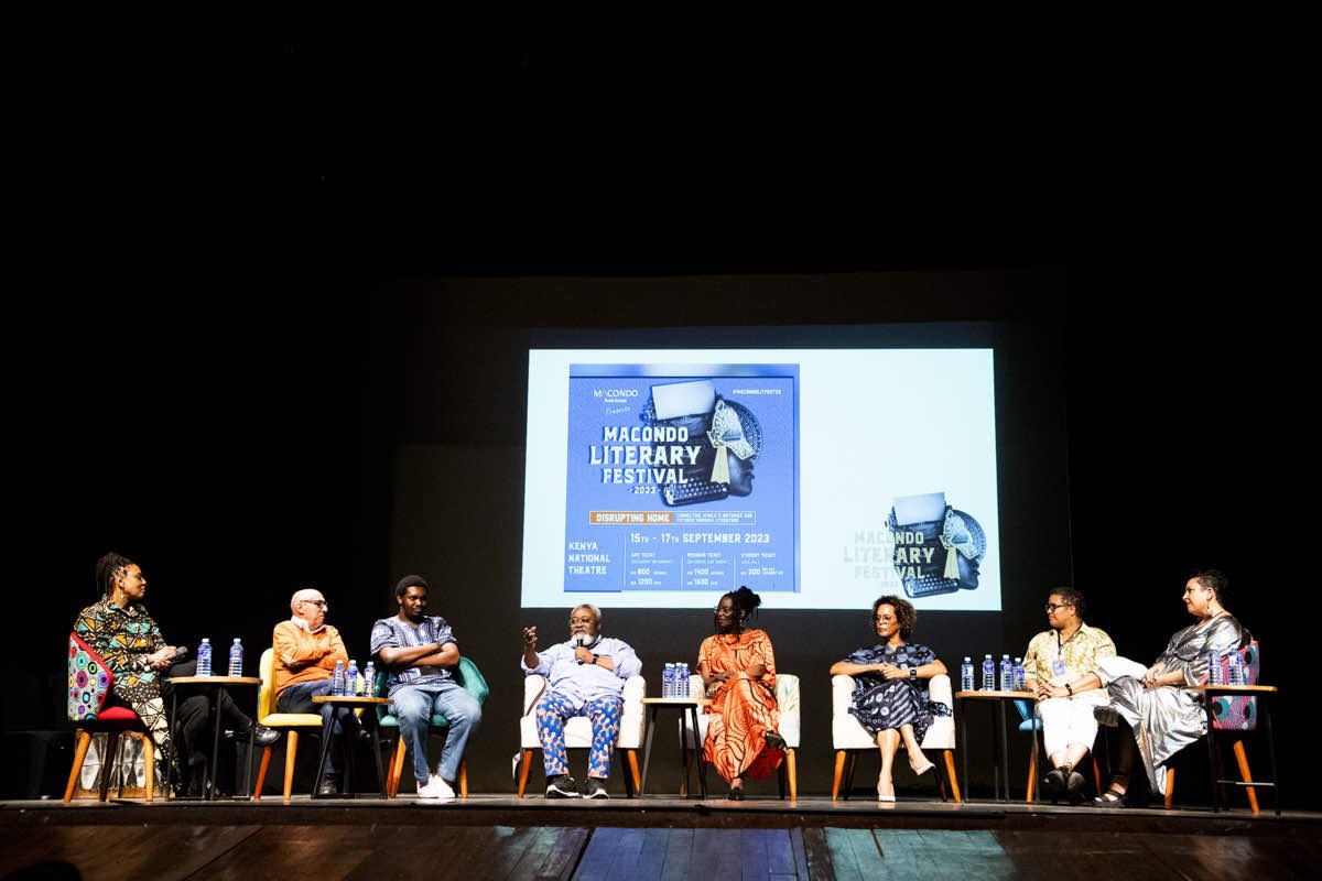 Panel discussion at the Macondo Literary Festival, with seven diverse speakers seated on stage, some holding microphones, with a large poster for the event projected in the background.