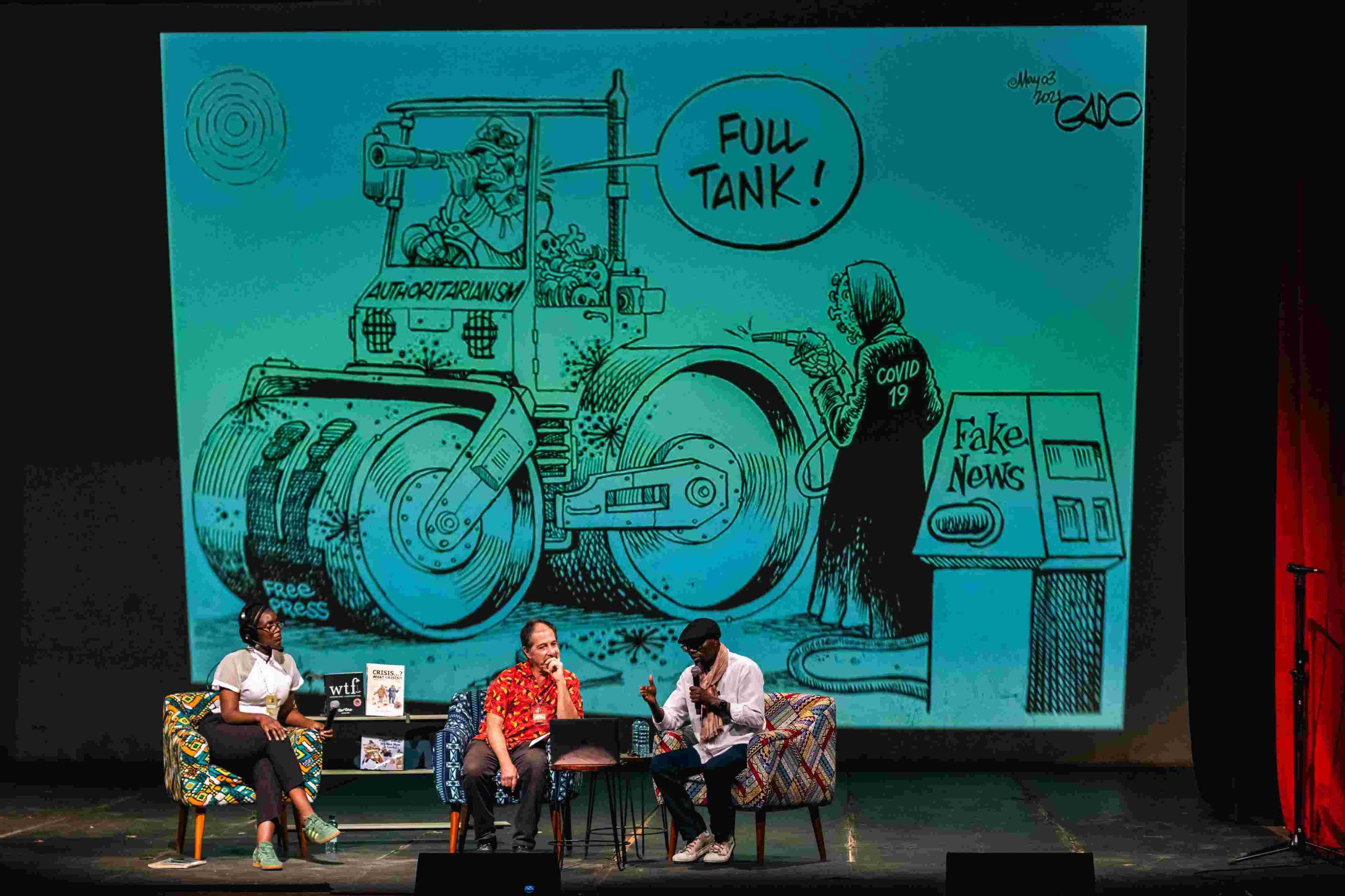 A panel discussion taking place on a stage with three people seated on colorful chairs, in front of a large cartoon-style poster. The poster depicts a person in a tank labeled "Autoritarianism," shooting at a woman with a speech bubble saying "Full T