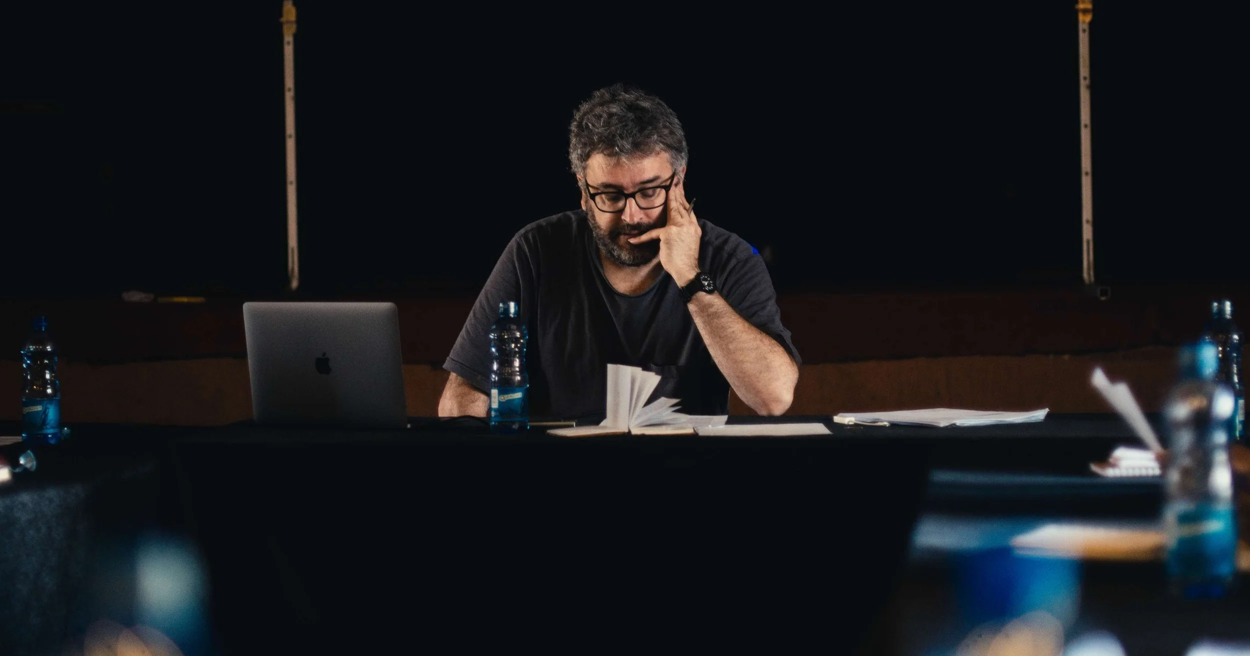 A man with glasses, a beard, and dark hair, seated at a table in a dark room, reading a book. There are water bottles, a laptop, and papers on the table.