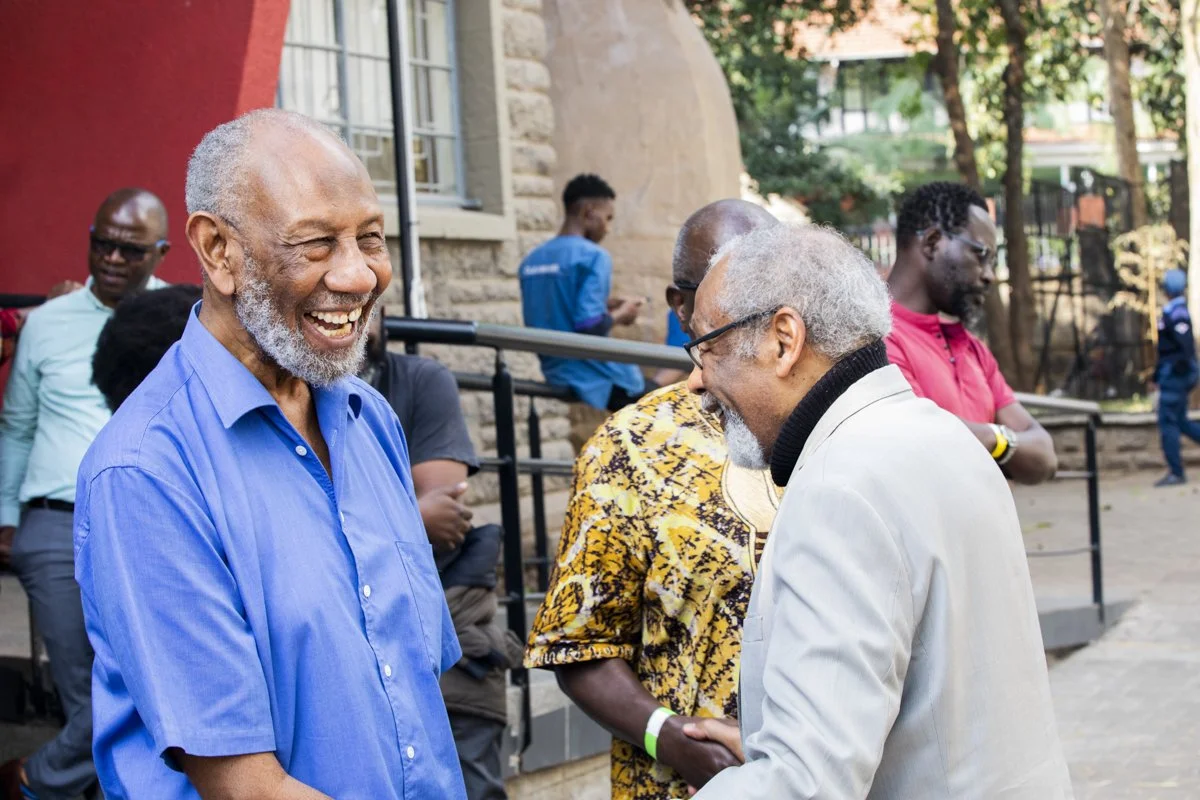 Two older men are smiling and engaging in a friendly conversation at an outdoor gathering, with other people in the background.