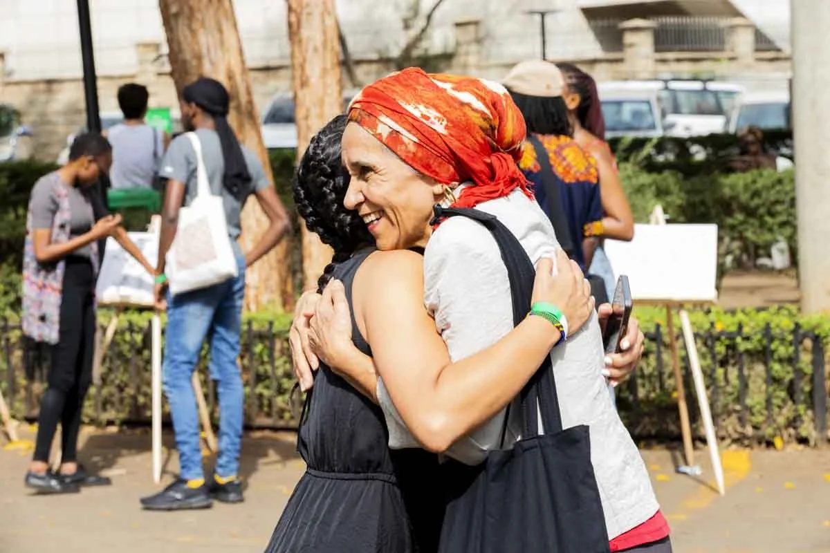 Two women hugging outdoors, smiling, one wearing a red headscarf. Several people are in the background, some looking at their phones, with trees and parked vehicles behind them.