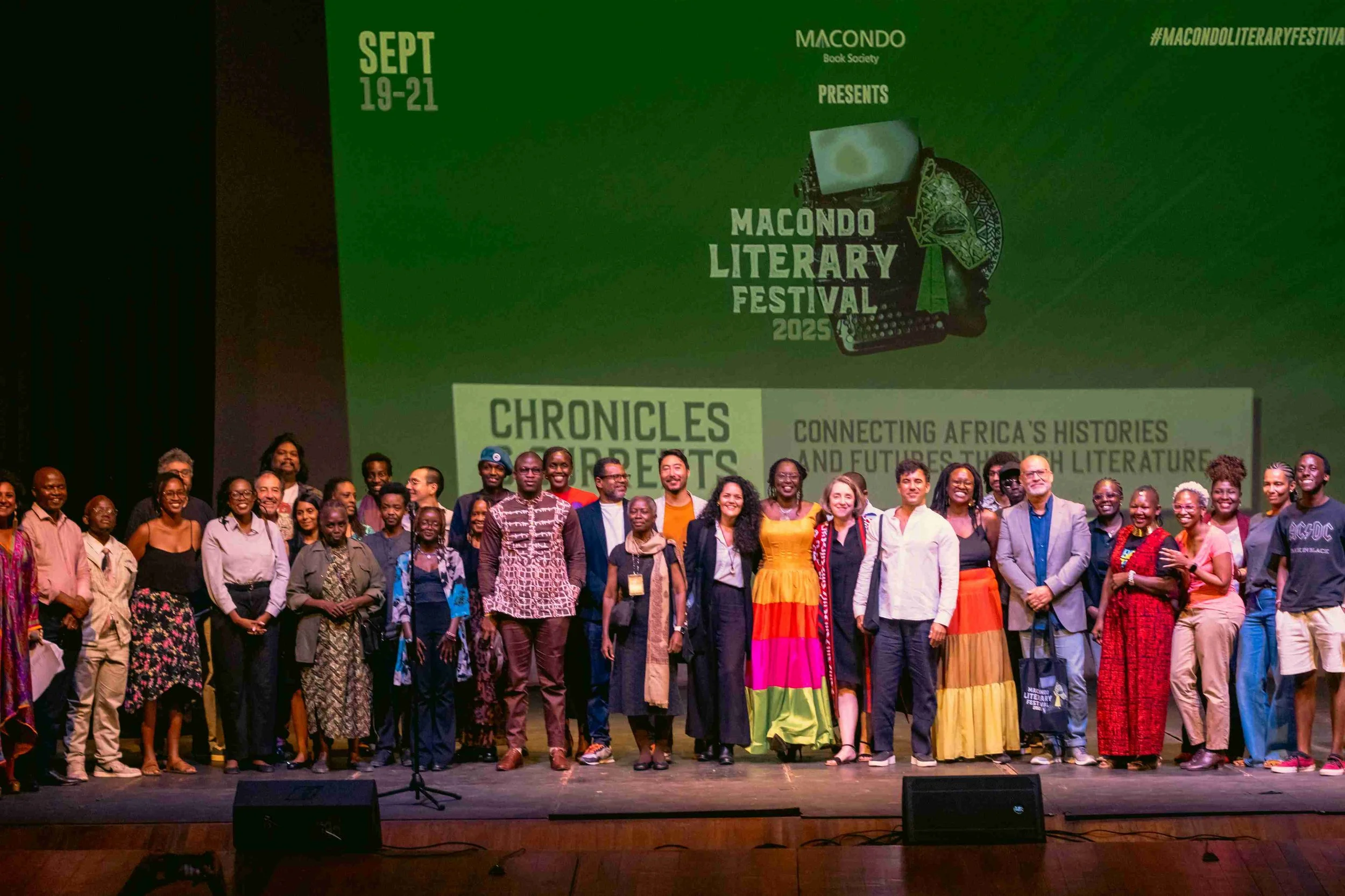Group of diverse people standing on stage at the Macondo Literary Festival 2025, with a large green screen behind them displaying event details and titled 'Chronicles and Reports'.