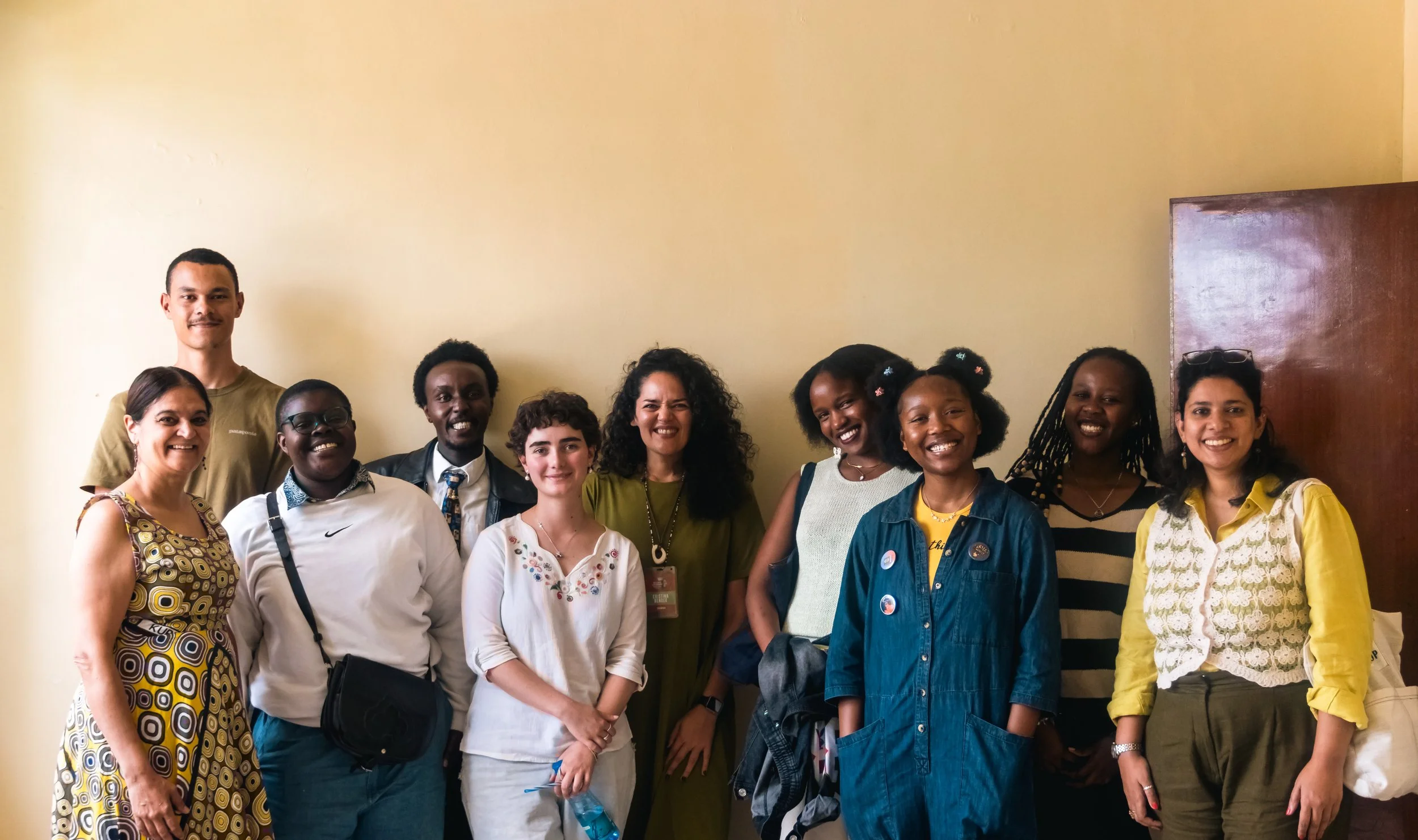 A diverse group of eleven people, including men and women of various ages and ethnicities, standing together indoors against a plain beige wall, smiling for the photo.