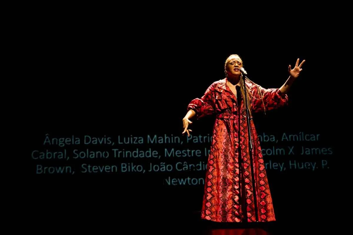 A woman in a red patterned dress singing into a microphone on stage with a black background. Names of performers are displayed below in blue text.
