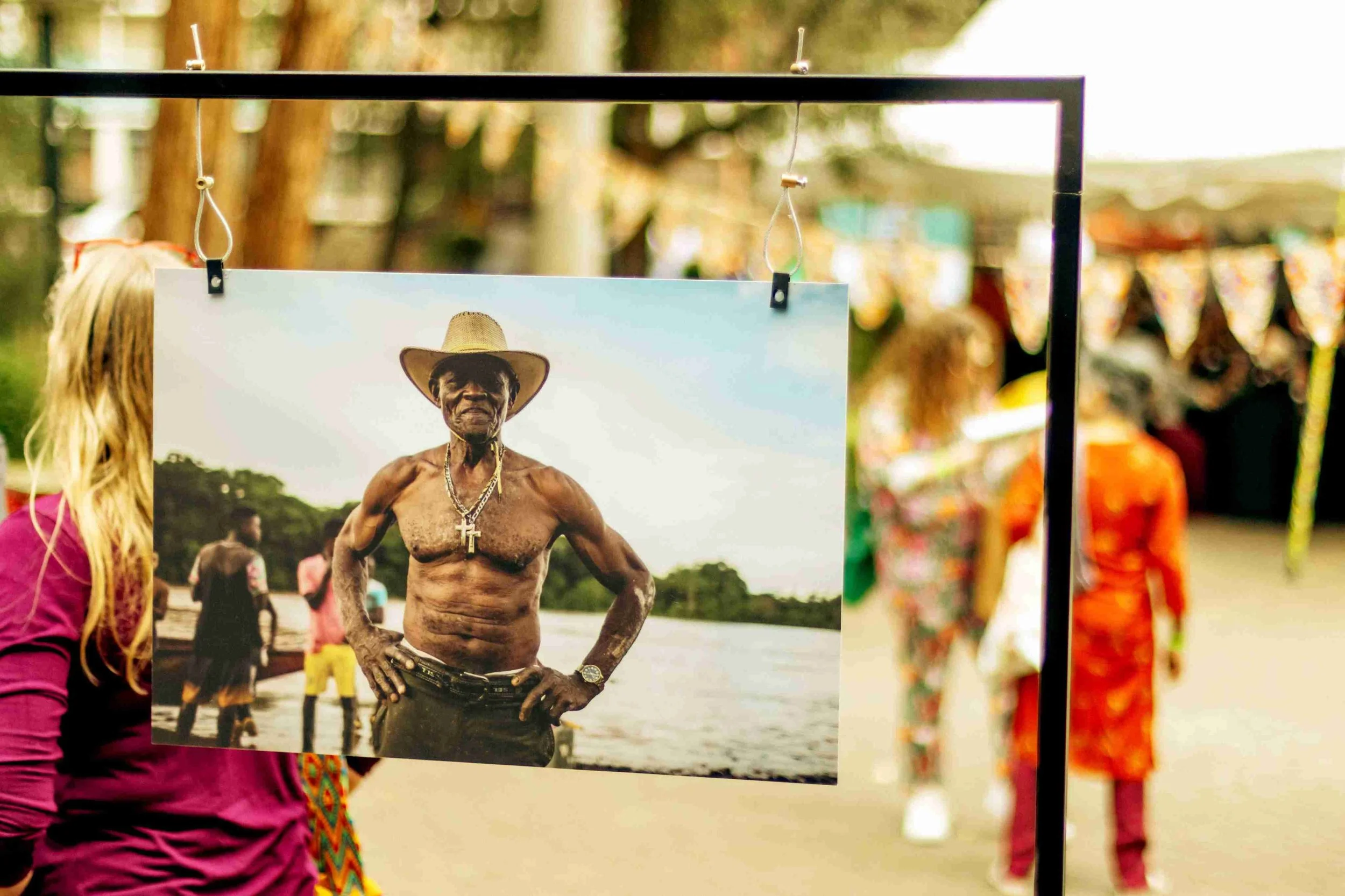 Photograph of an elderly shirtless man wearing a cowboy hat and jewelry, standing outdoors near a body of water with trees in the background, displayed on a photo stand at an outdoor market or exhibition.
