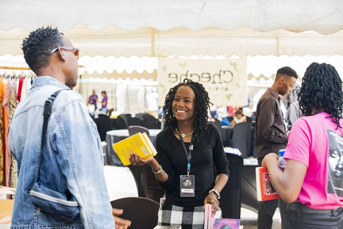 Three women and a man in a marketplace or trade fair setting, engaging in conversation. The woman in the center is smiling and holding a yellow book, wearing a black shirt and a conference badge, while the woman on the right, wearing a pink shirt, ho