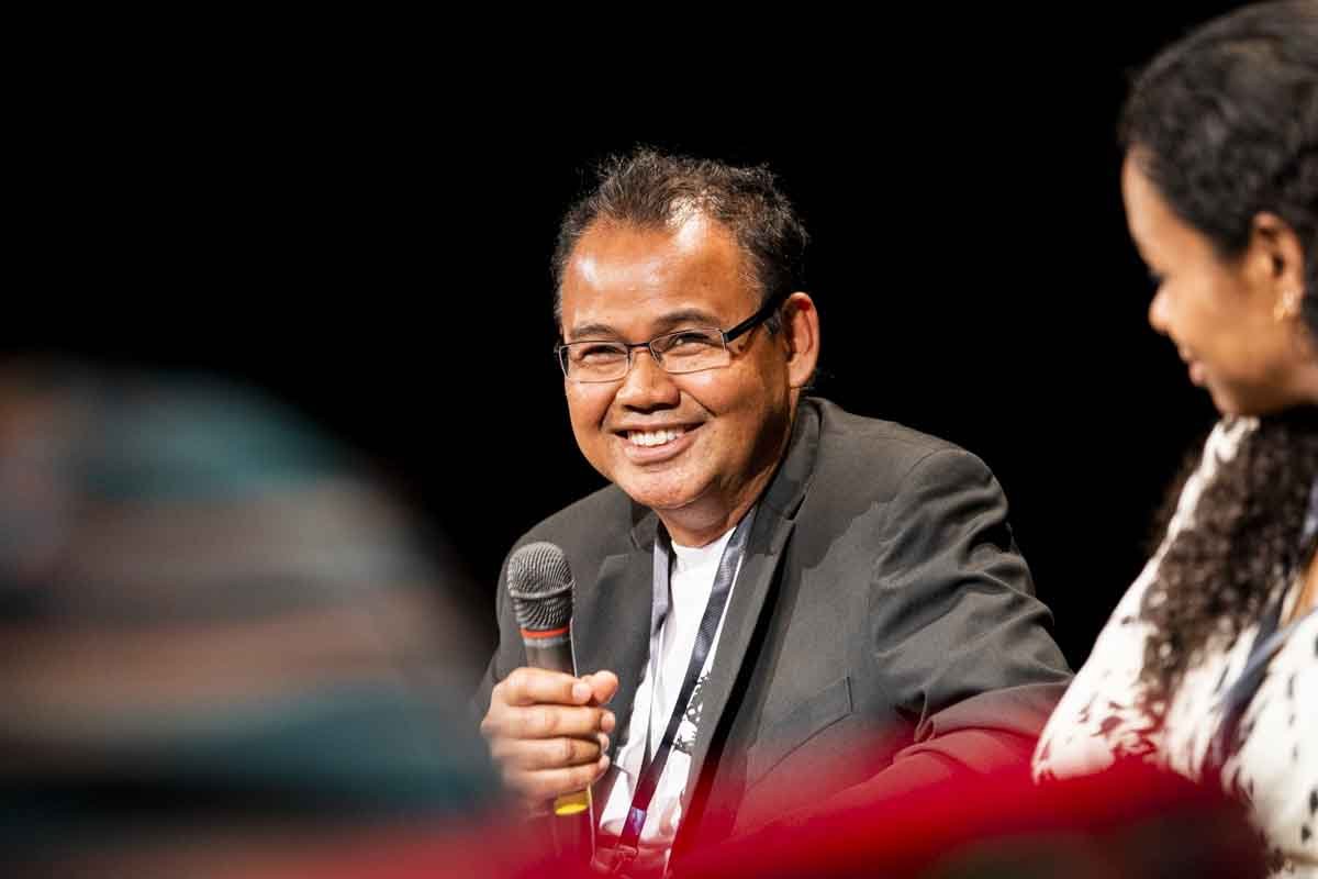 A man wearing glasses and a gray blazer smiling and holding a microphone, sitting next to a woman with dark hair in a patterned top during a panel discussion or interview, with a dark background.