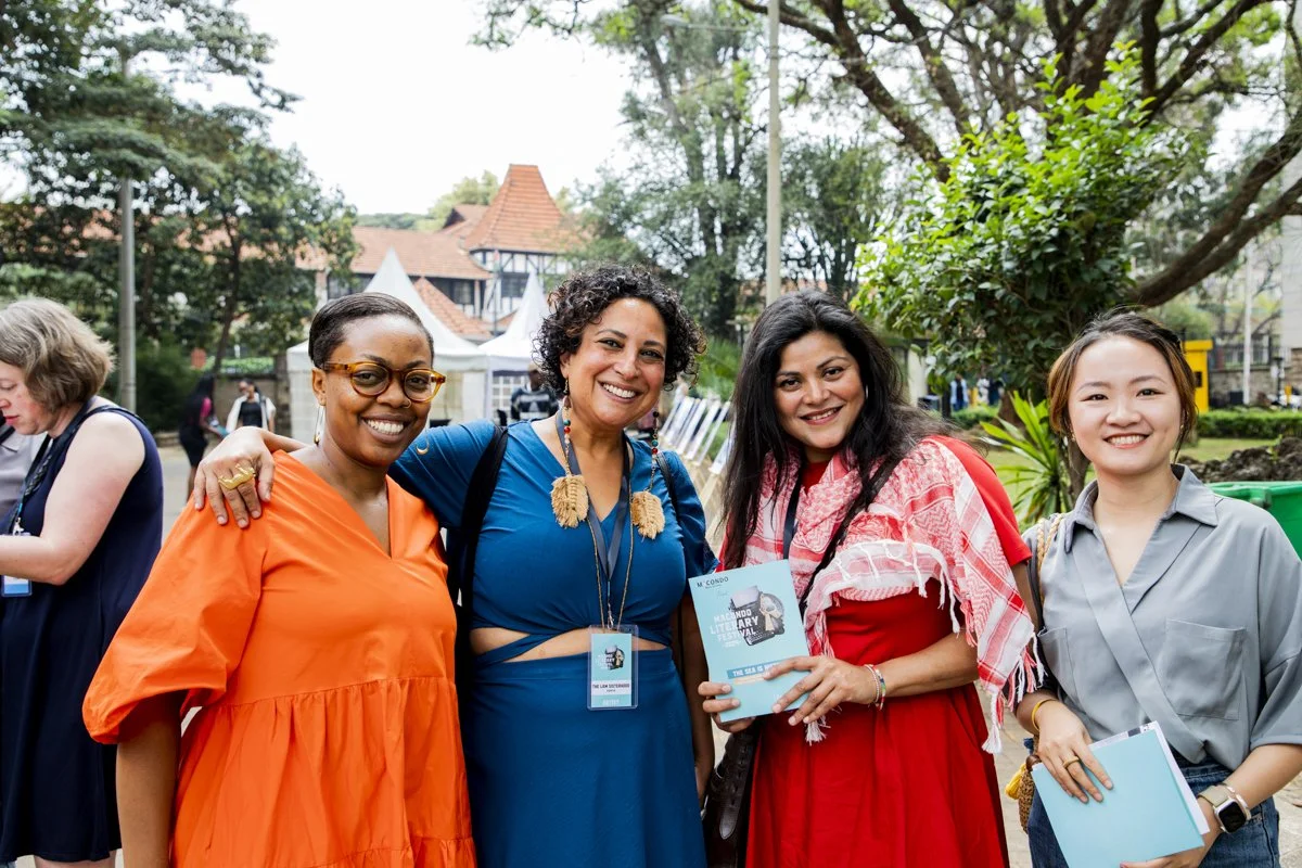 Four women smiling and posing outdoors at a park or garden, with trees and buildings in the background. One woman is holding a book.
