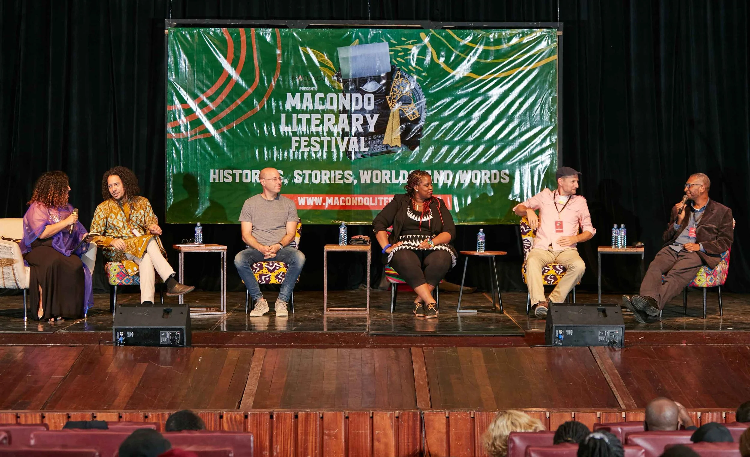 Six individuals participate in a panel discussion on stage at the Macondo Literary Festival. They are seated in a row, with a large green festival banner behind them that reads "Macondo Literary Festival - Stories, Words, World." Each person has a sm