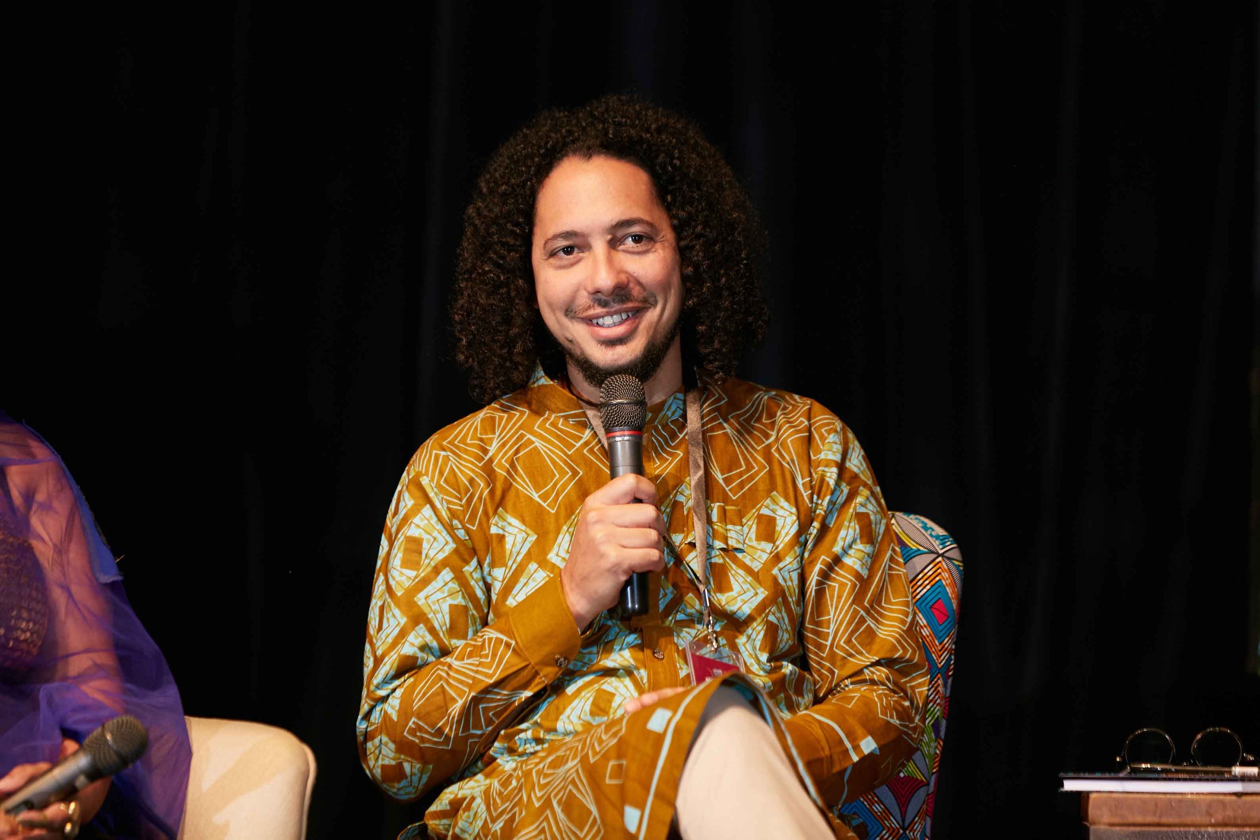 A man with curly hair and a beard smiles while holding a microphone, sitting on a stage with a dark background, wearing a patterned shirt.