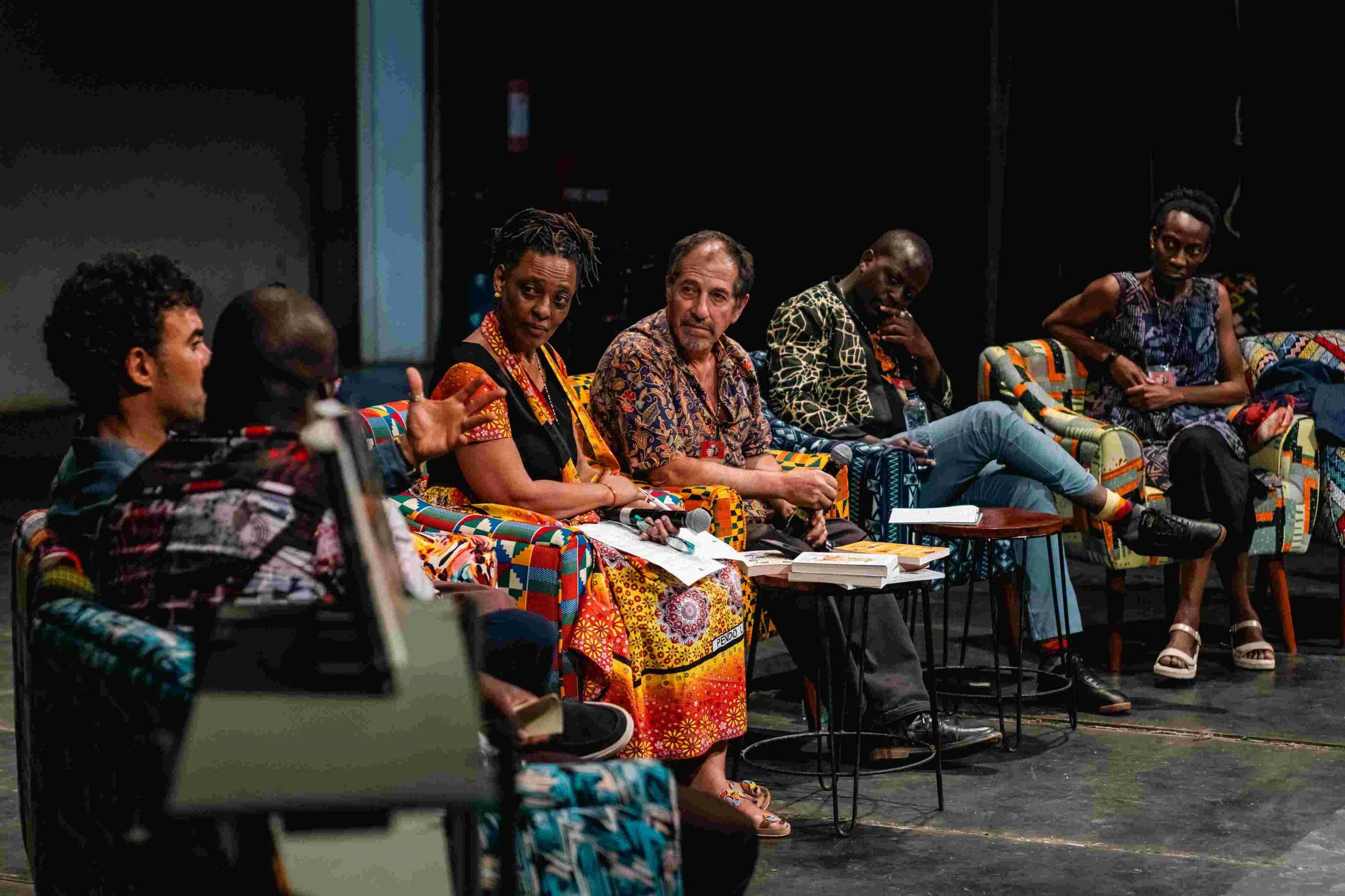 Panel of six people sitting on colorful chairs engaged in a discussion in a dark room, with books and microphones on small tables in front of them.