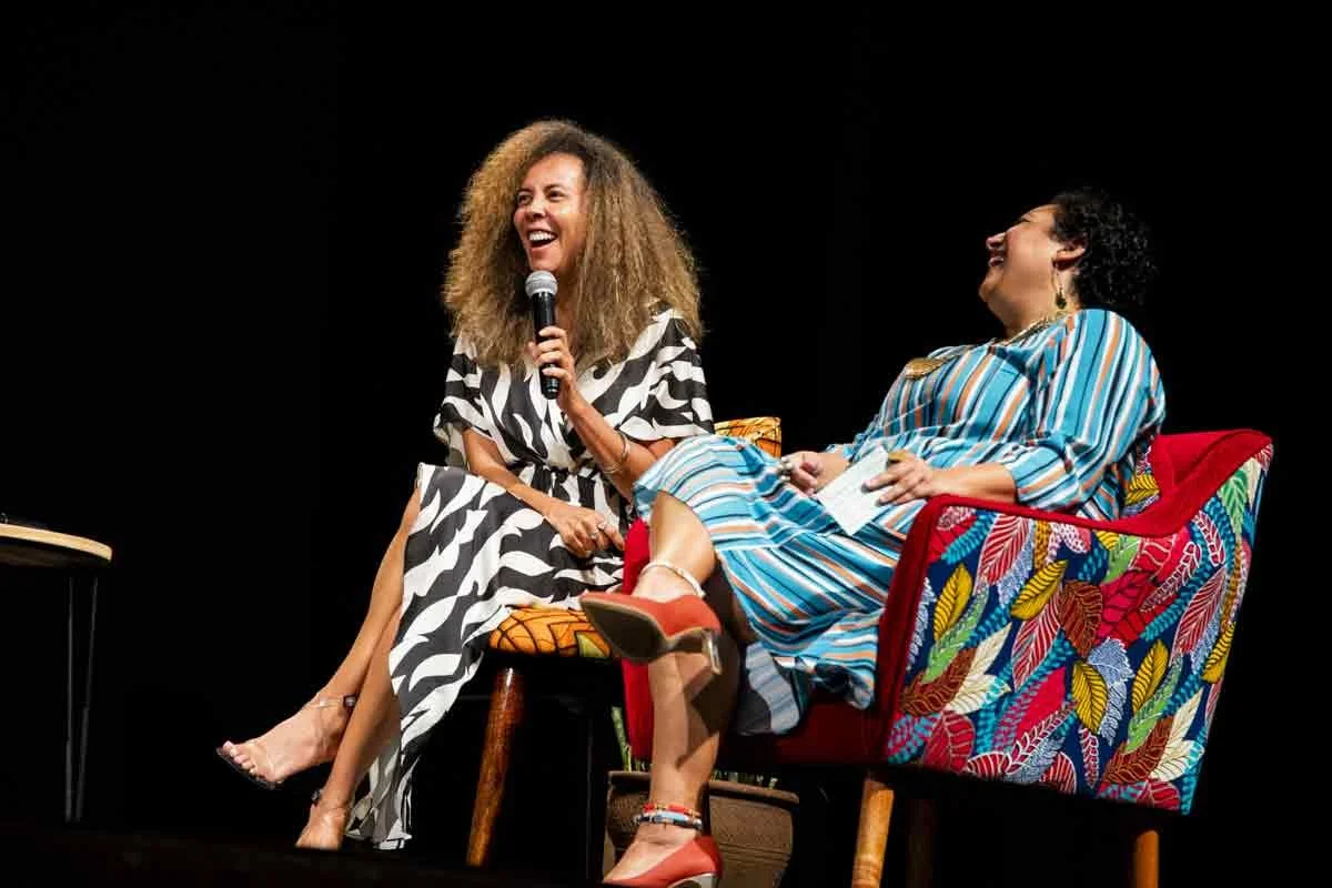 Two women are on stage, one holding a microphone and laughing, the other sitting on a colorful armchair, also laughing. They are engaged in a joyful conversation.