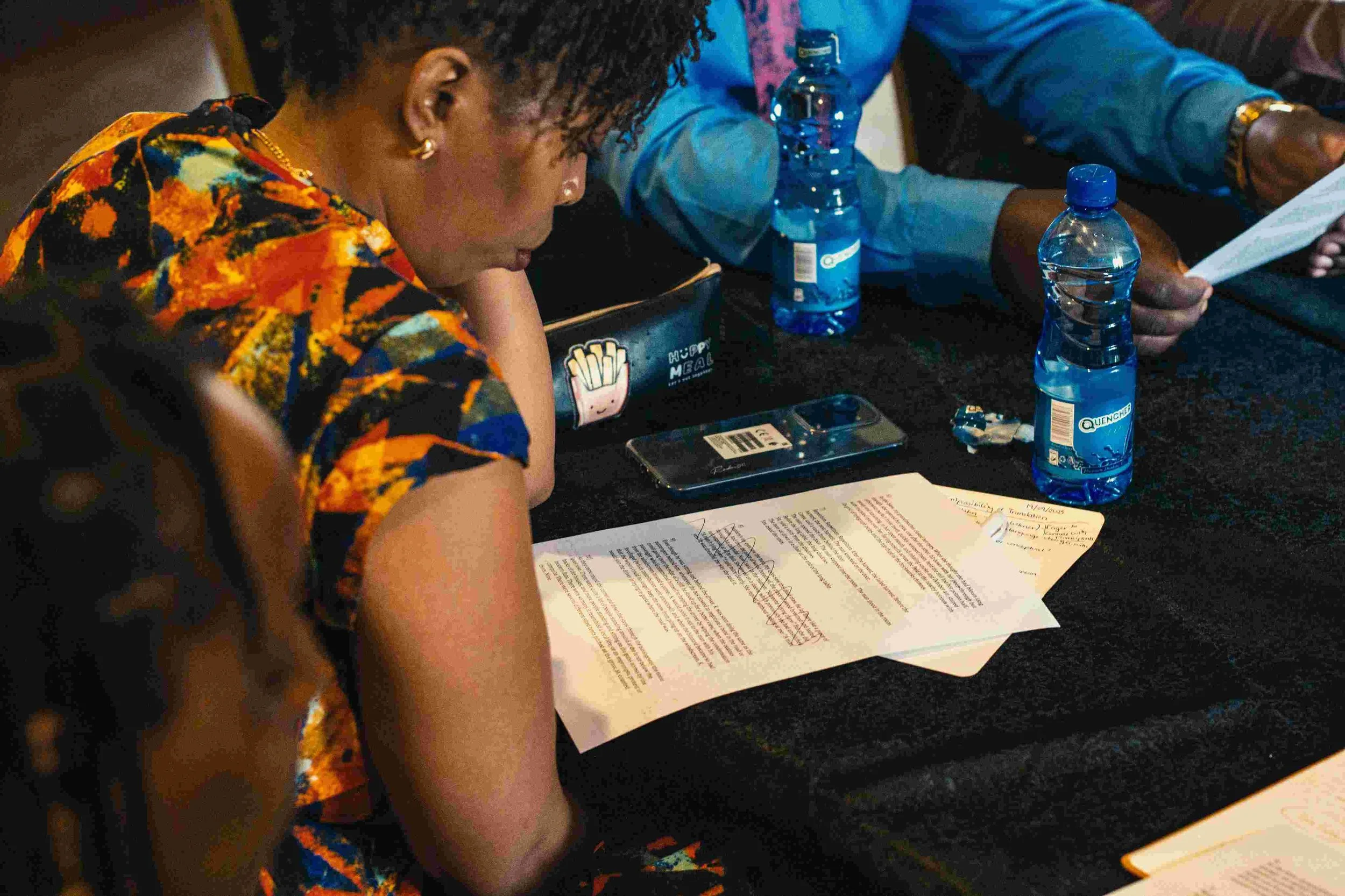 A woman with curly hair wearing a colorful patterned dress sits at a table, reading a document, with several bottles of water, a phone, and a purse on the table.