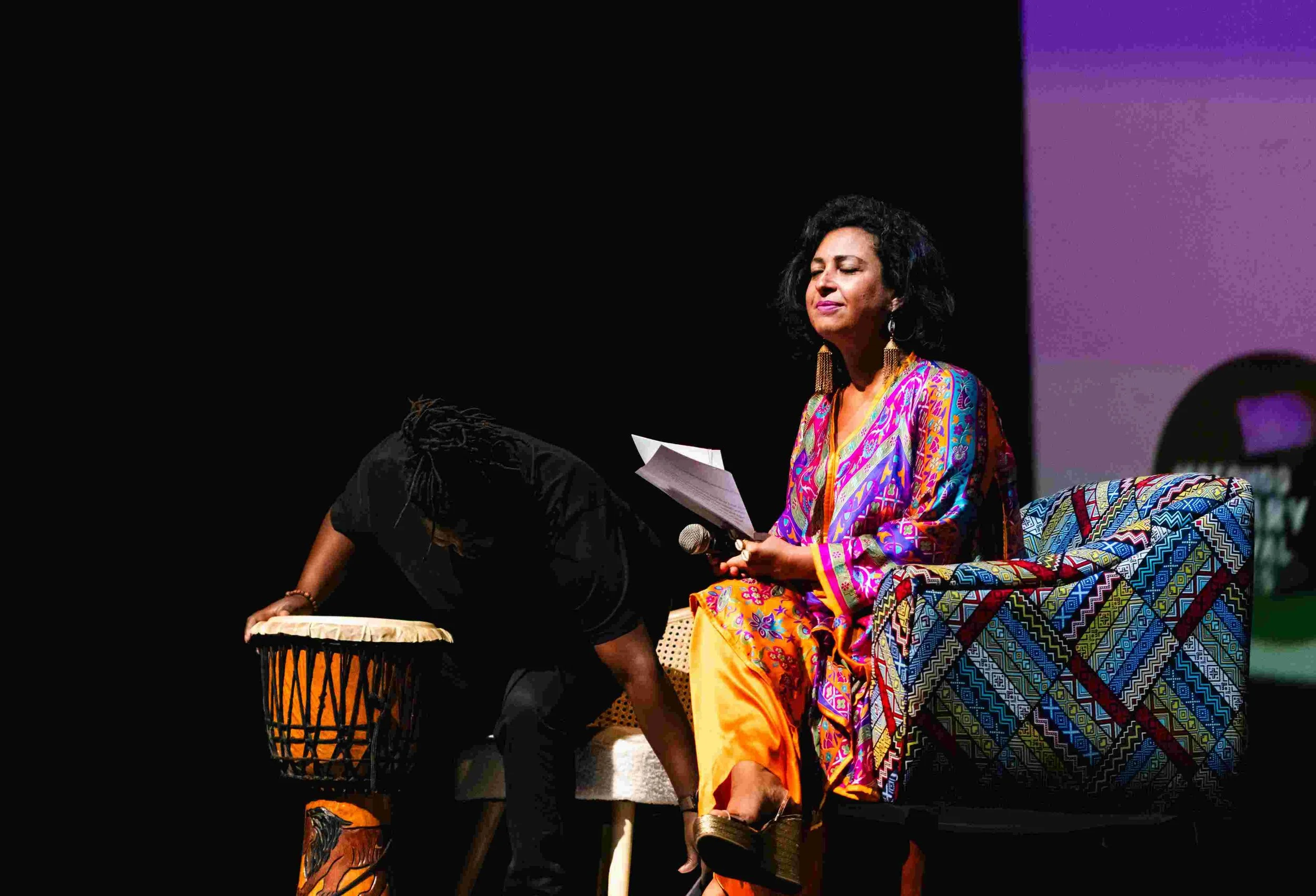 A woman in colorful traditional attire sitting with eyes closed, holding papers and a microphone, next to another person bending over a drum on stage.
