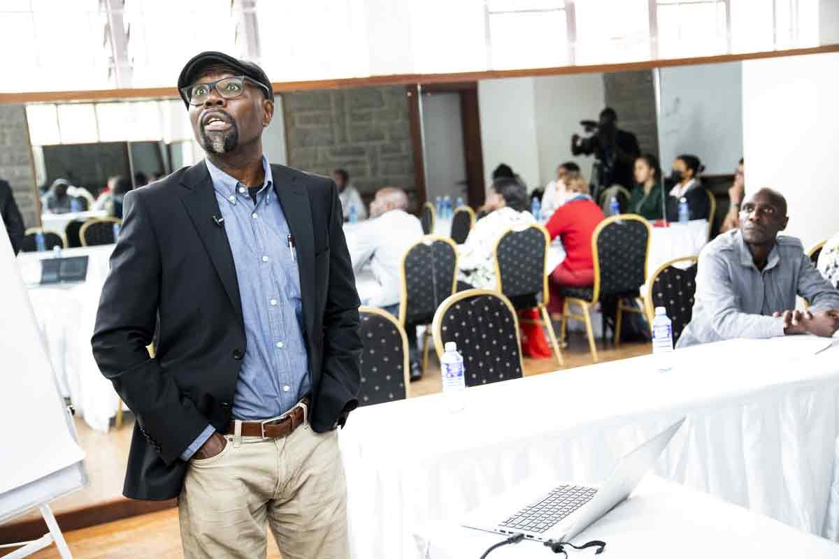 Man wearing glasses, a cap, a black blazer, a blue shirt, and khaki pants stands at a presentation or conference, with a group of seated attendees in the background.