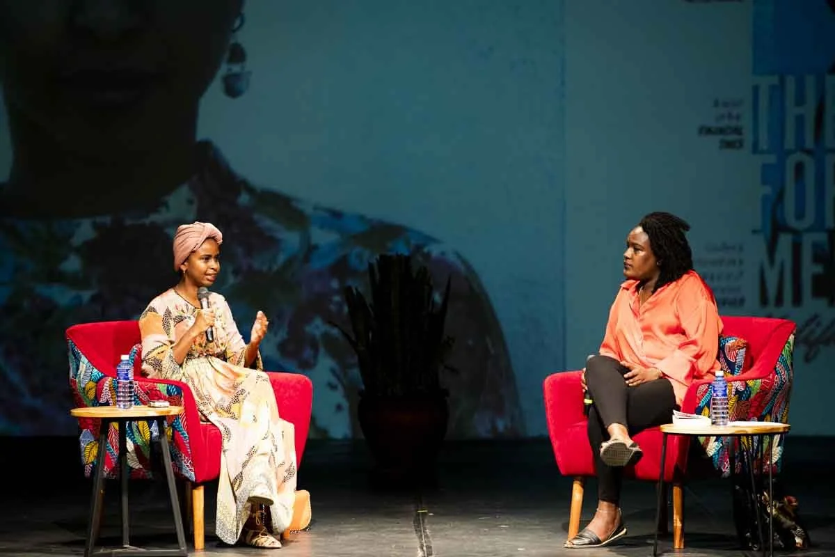 Two women sitting in red chairs on a stage engaged in conversation, each with bottled water on a side table. One woman wears a beige and patterned dress with a head wrap, and the other wears a peach blouse and black pants.