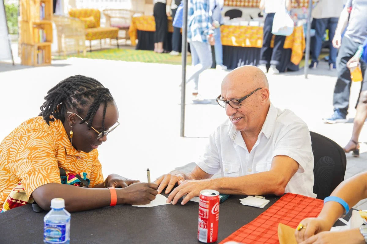 A man with glasses and a white shirt sits at a table with a young woman with braided hair, sunglasses, and a colorful orange shirt. They are signing a paper, and there are items on the table including a water bottle, a can, and some papers. The setti