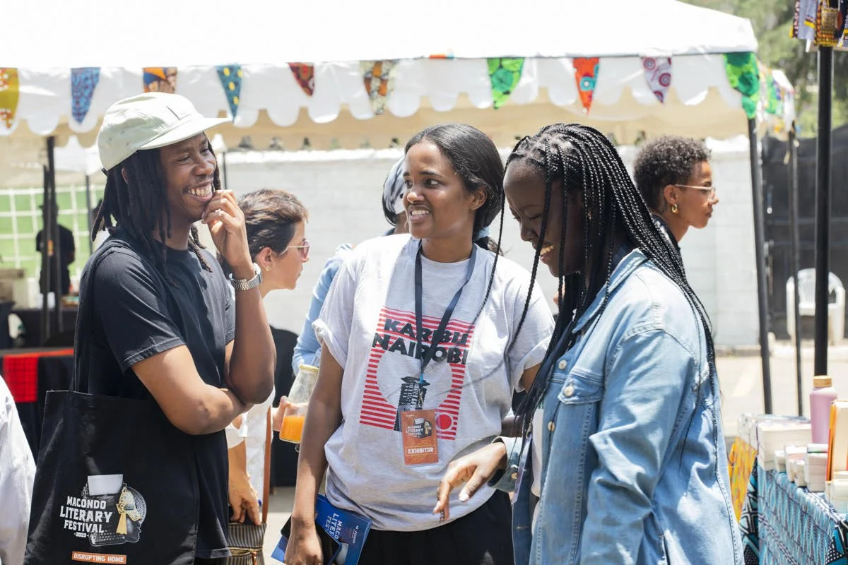 Group of women at an outdoor festival, laughing and chatting under a decorated canopy, with books and bottles on a table nearby.