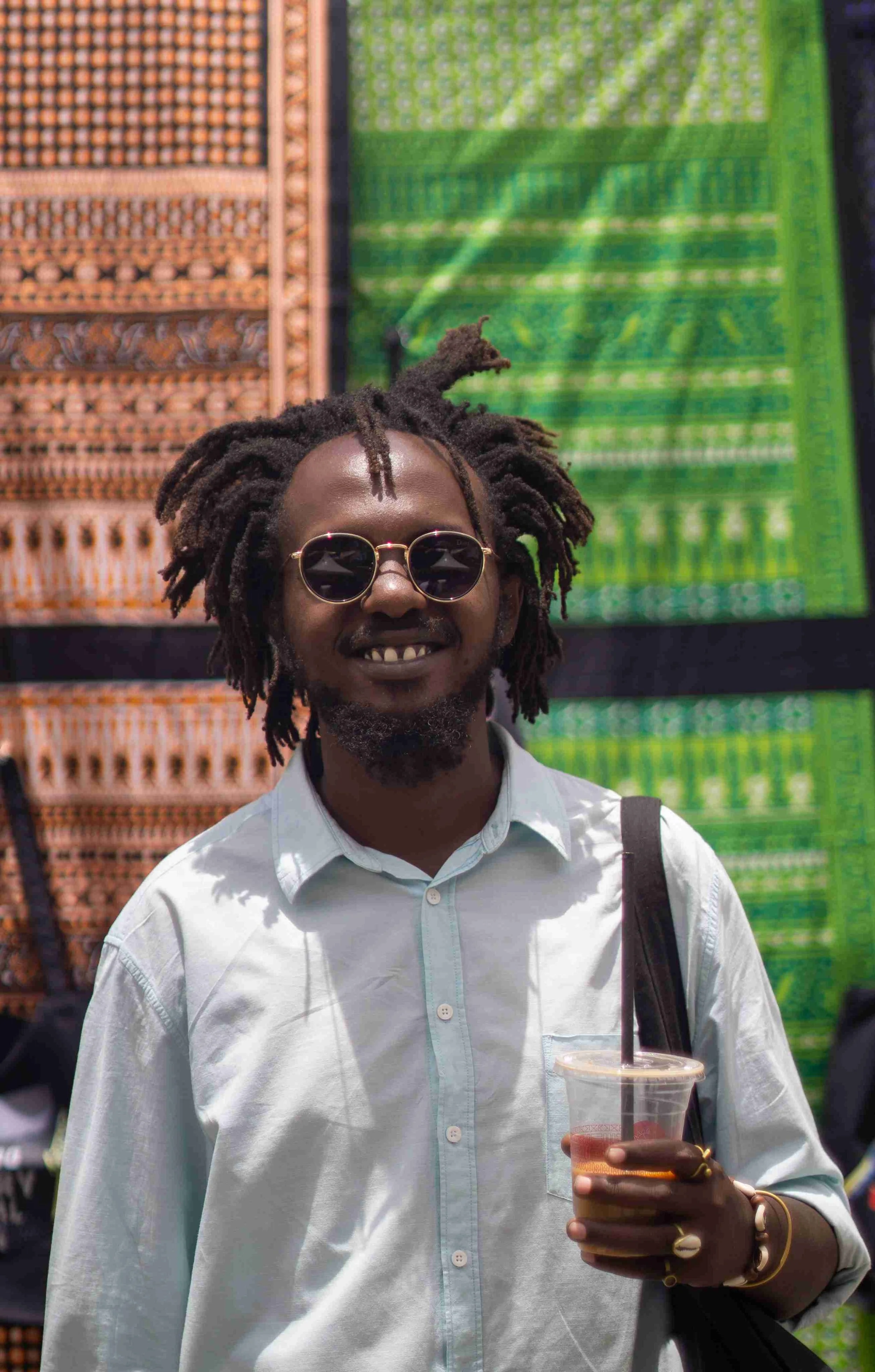 A man with dreadlocks, wearing sunglasses and a white button-up shirt, holding a plastic cup with a drink, smiling at an outdoor event with colorful woven textiles in the background.