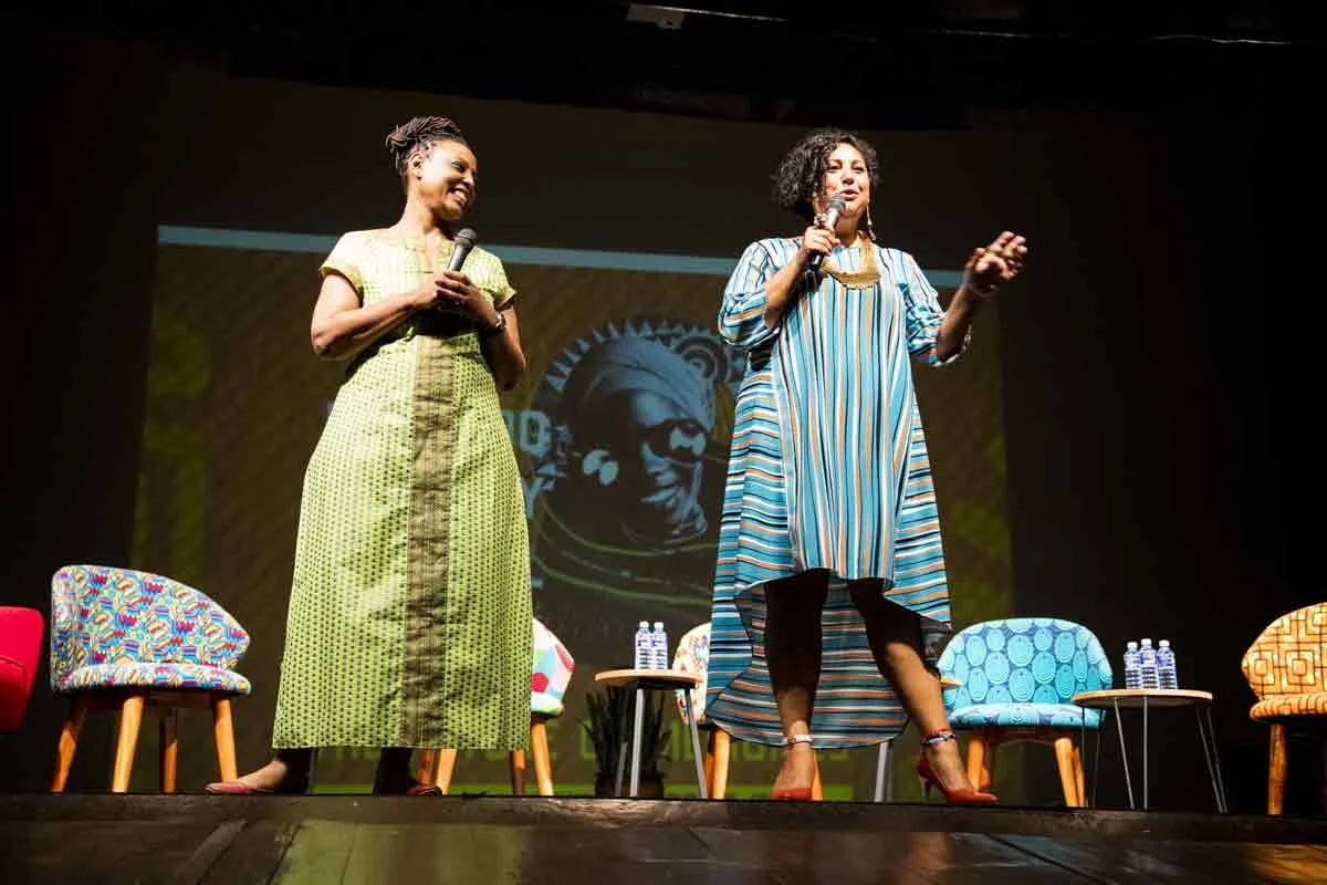 Two women speaking on stage with microphones, seated on colorful chairs, and a large screen behind them showing a logo of a face with glasses.