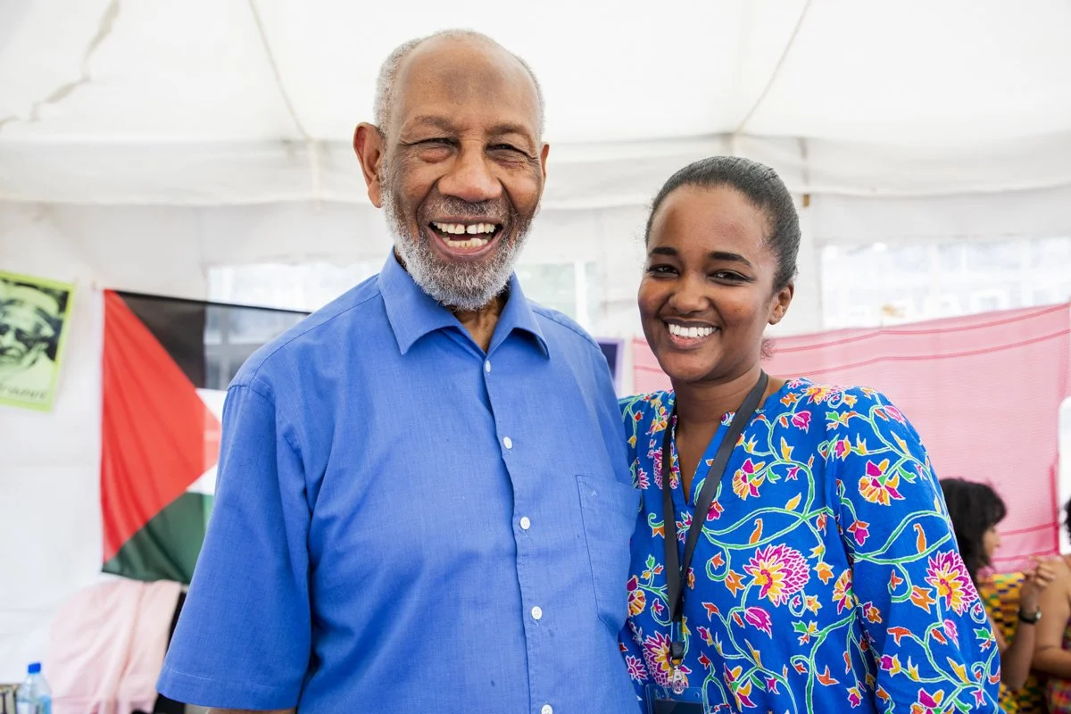 An elderly man with gray hair and beard wearing a blue shirt, smiling. A young woman with dark hair in a bun, wearing a colorful floral top and a lanyard, smiling. They are indoors with a white tent and some flags or banners in the background.