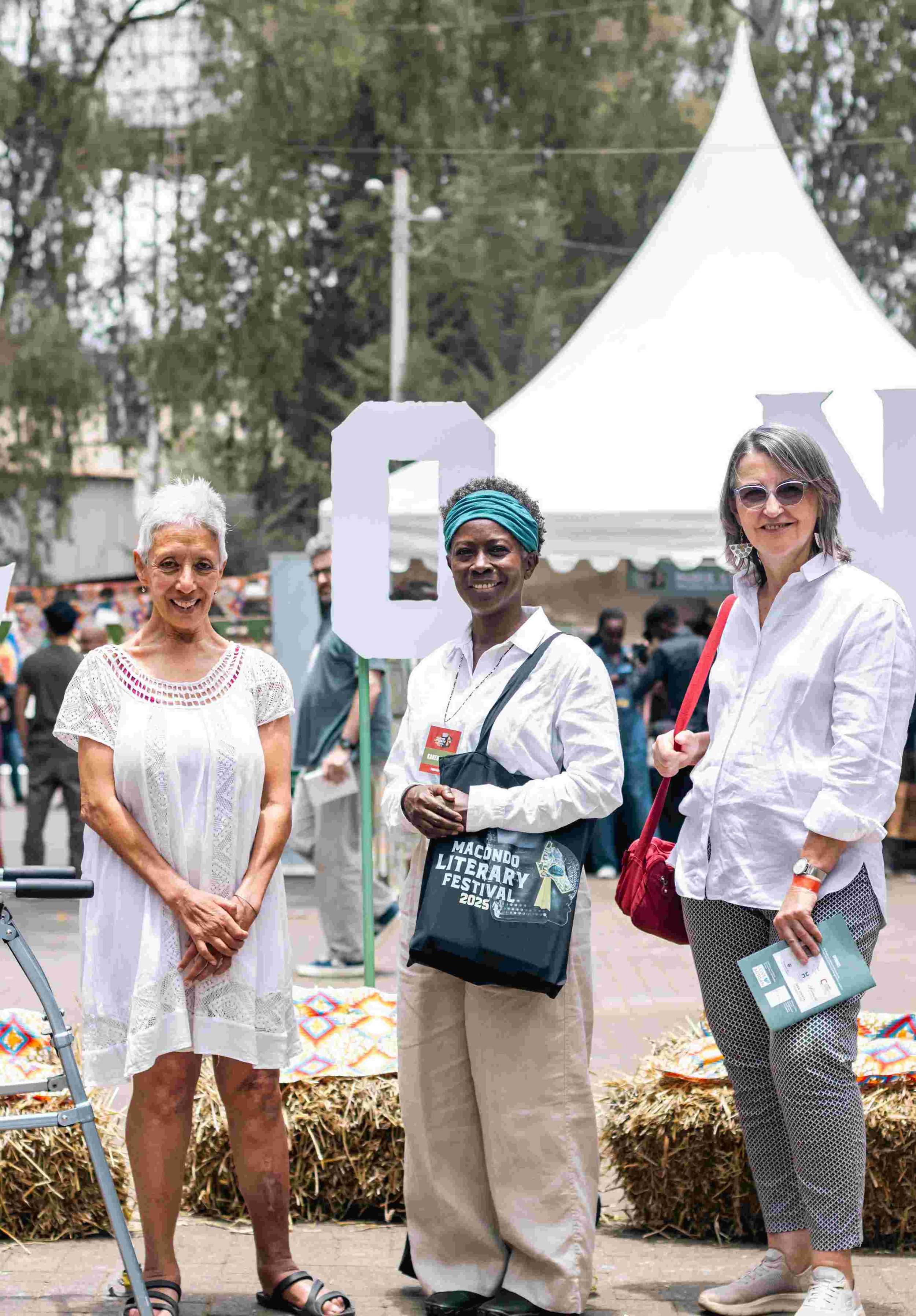Three women smiling at an outdoor event, standing in front of a white tent. One holds a brochure, another has a tote bag with 'Macondo Literary Festival 2023' on it, and the third woman wears sunglasses and a white shirt. There are other people and e