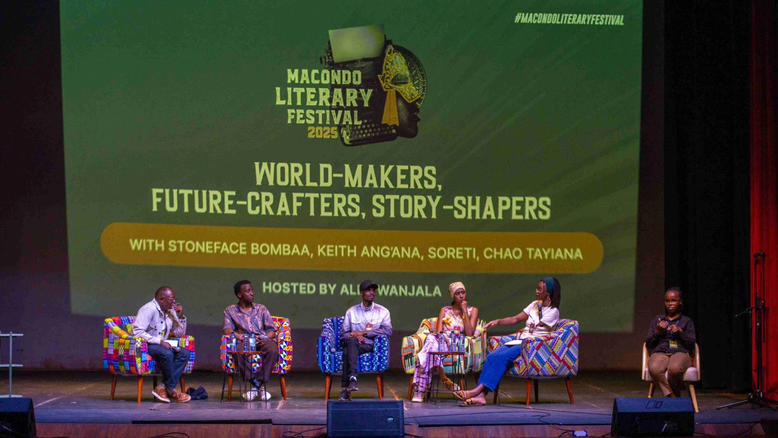Panel discussion at Macondo Literary Festival 2025 with six speakers seated on stage in colorful chairs, titled 'World-Makers, Future-Crafters, Story-Shapers' with hosts and guest speakers' names displayed on a large screen behind them.
