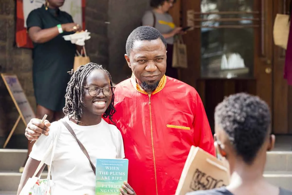 A woman with glasses and a white shirt stands next to a man in a red jacket, both smiling and posing for a photo in a busy outdoor setting. There are two other people in the background, one on the phone and another holding a tray and a paper bag.