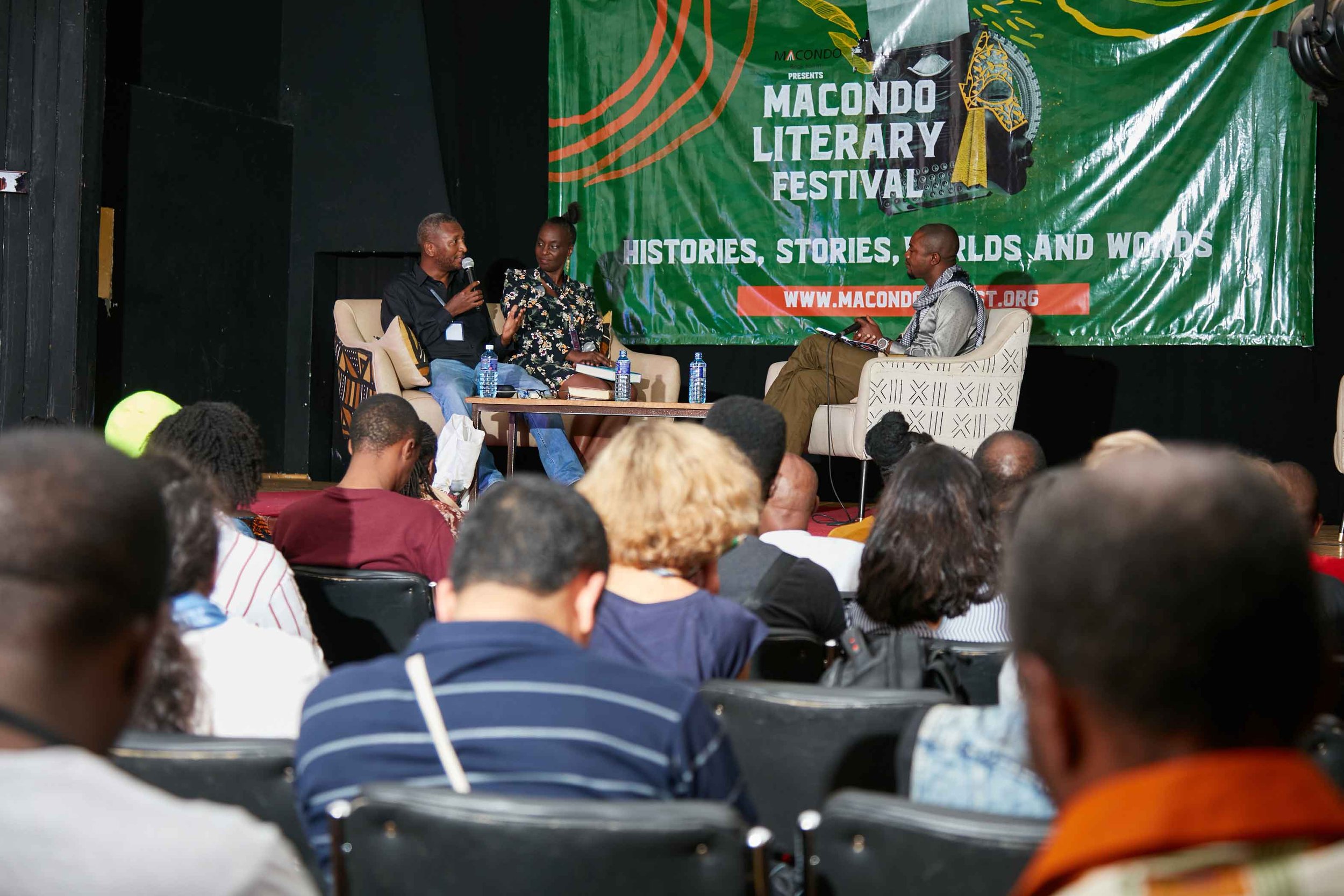 Panel discussion at the Macondo Literary Festival with three speakers on stage, two sitting on beige armchairs and one on an office chair, with a large green banner in the background promoting the event and featuring the text 'Macondo Literary Festiv